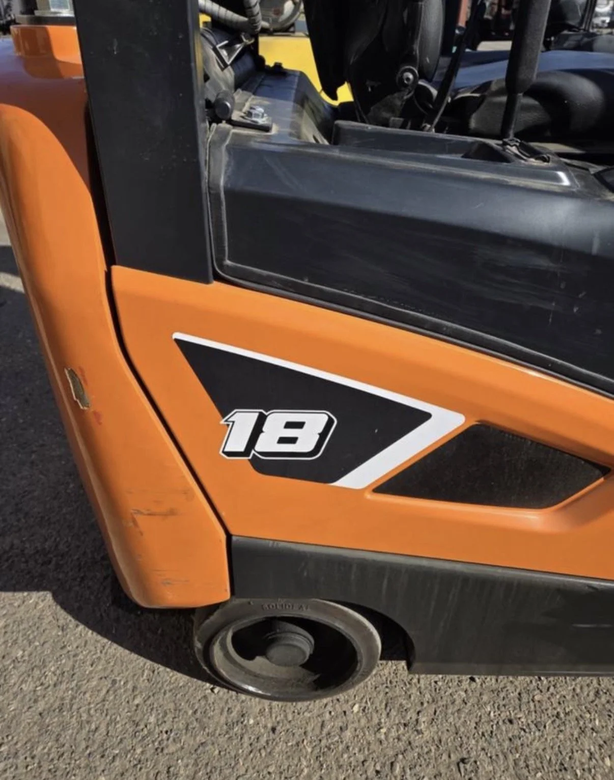 Close-up of an orange race car with a black and white number 18 decal on its side, showing part of the front wheel, tire, and black bumper.