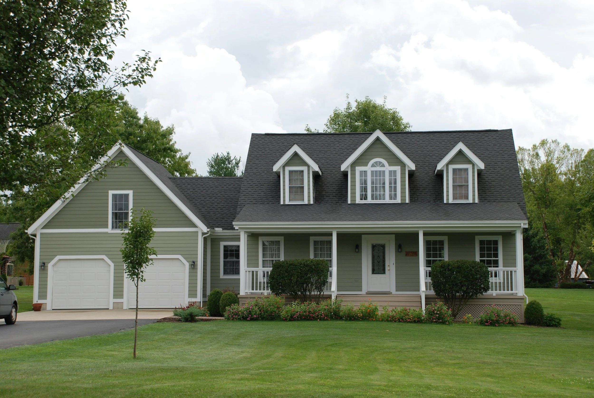 A two-story house with light green exterior, dark shingle roof, and white trim, featuring dormer windows on the upper level. The front porch has white railings and is decorated with bushes and flowers. There is a two-car garage on the left side and a driveway with a car partially visible. The house is surrounded by a well-maintained lawn and trees.