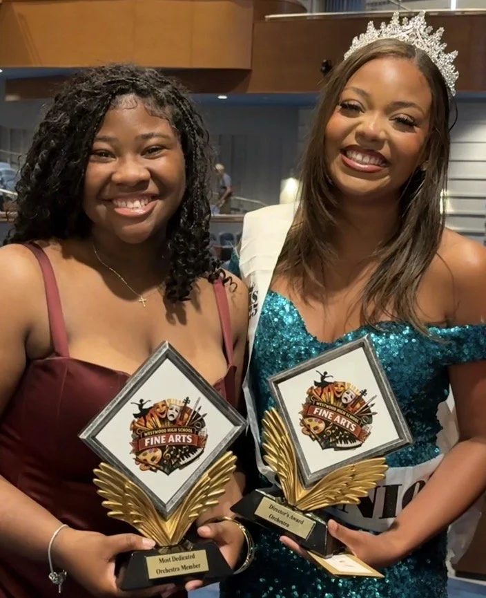 Two women smiling, holding trophies, one with a tiara and a sash, indicating a pageant or competition celebration.