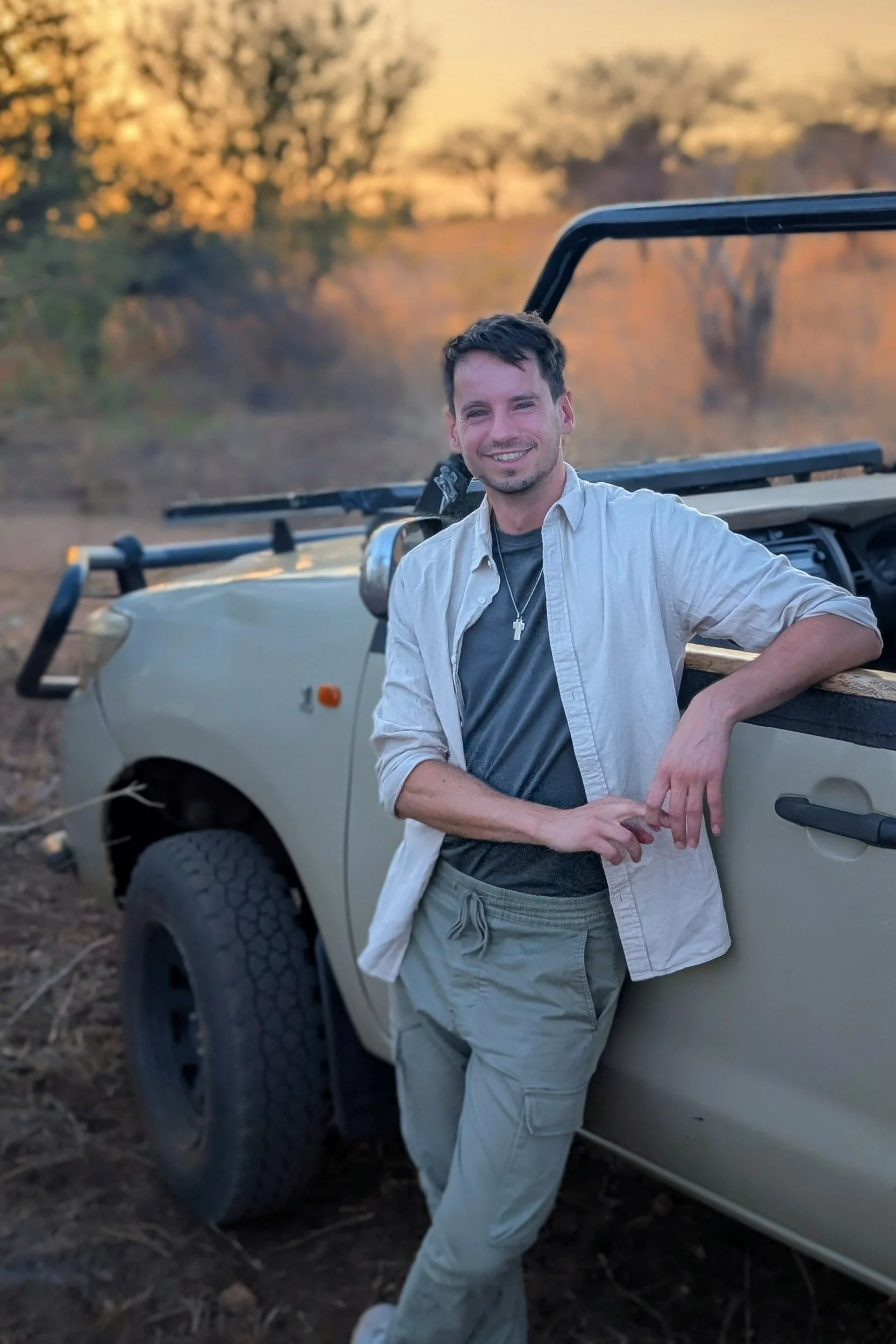 A young man with short dark hair, smiling, wearing a light-colored button-up shirt over a darker shirt, standing next to a beige off-road vehicle in a rural outdoor setting during sunset or late afternoon, with trees and warm lighting in the background.