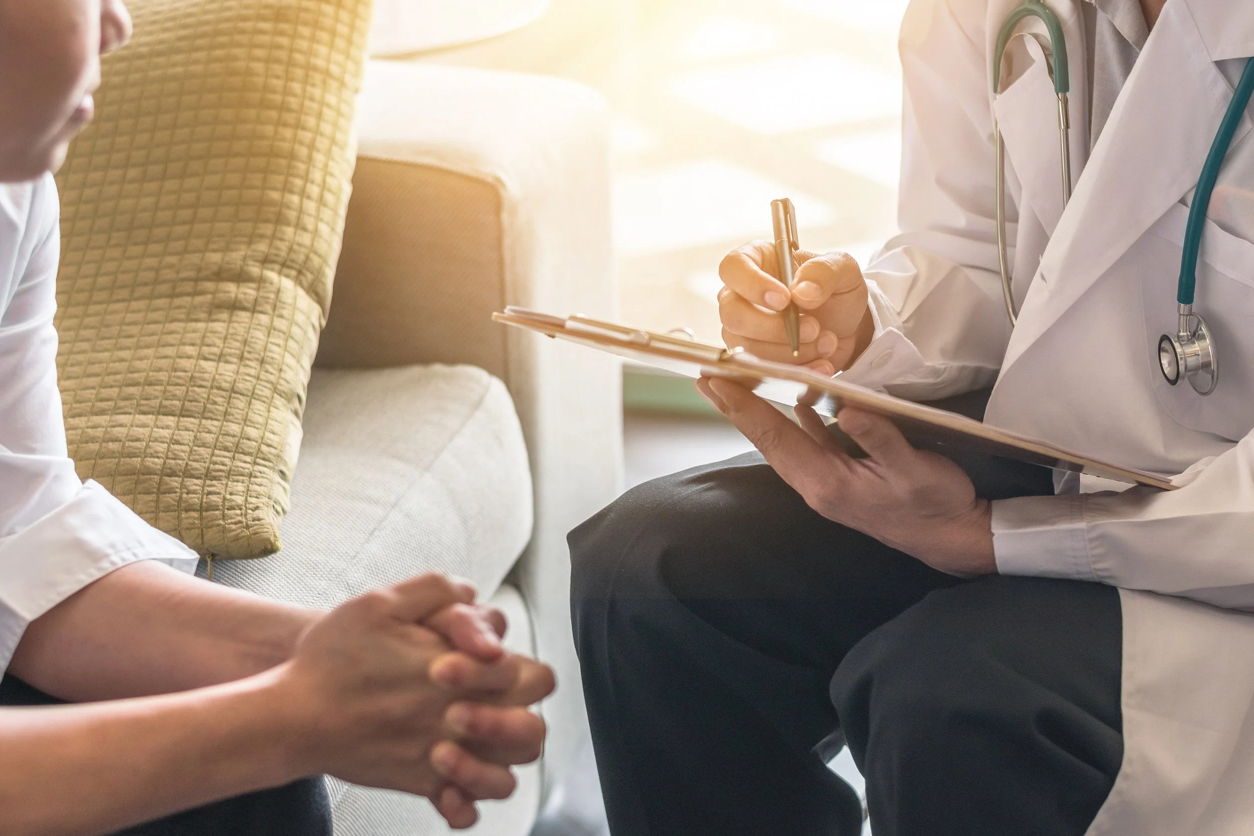 Clinician in a white coat taking notes while a patient sits on a sofa.