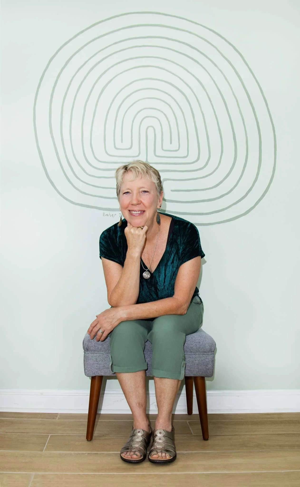 Lisa Garret, owner of Labyrinth Colon Hydrotherapy, shown sitting and smiling, against a pale green wall with a mural of a labyrinth on it.