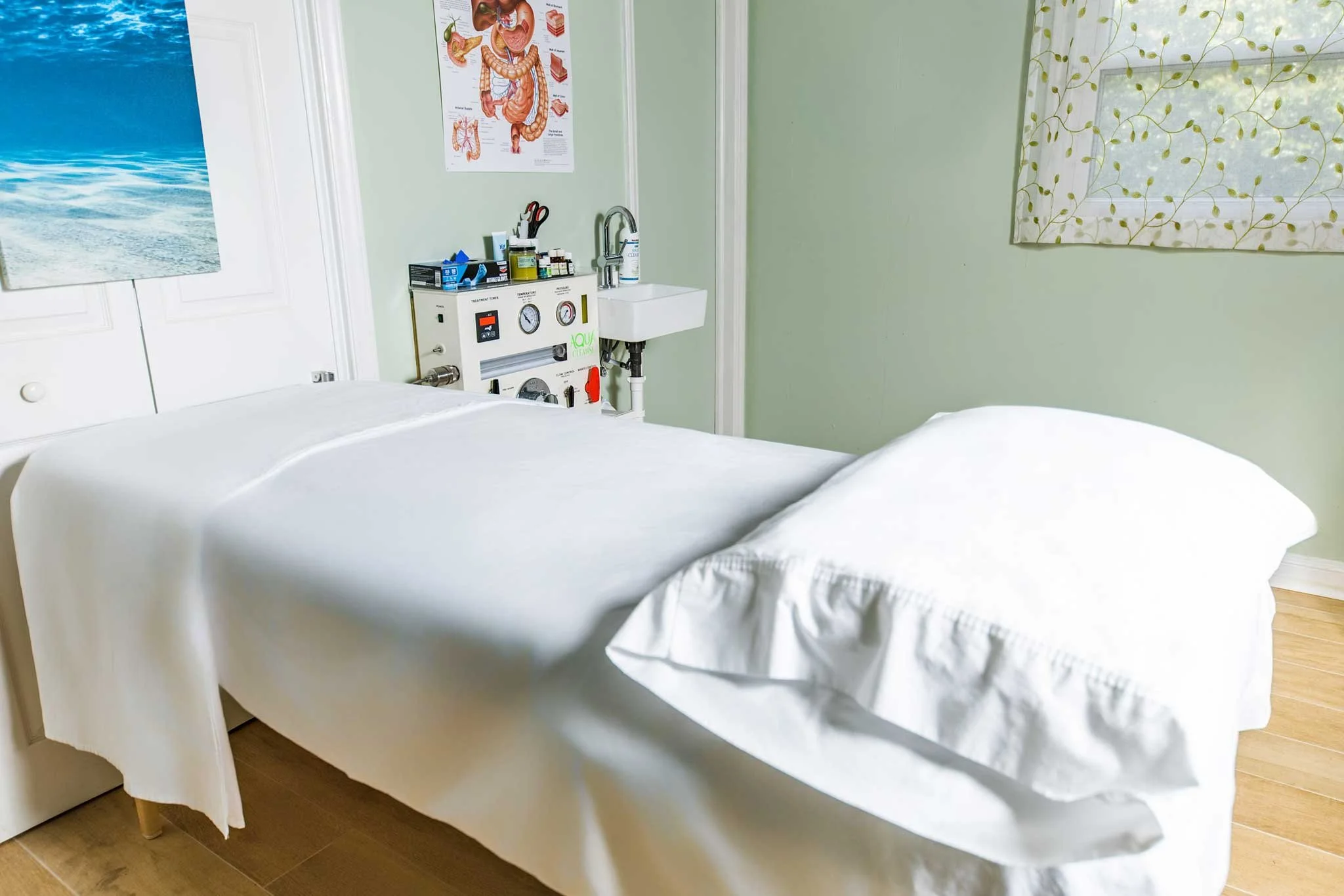 A treatment room with an unmade bed covered in white sheets, medical equipment stand with supplies nearby, and a window with a leaf-patterned curtain. A poster of the human digestive system is on the wall.