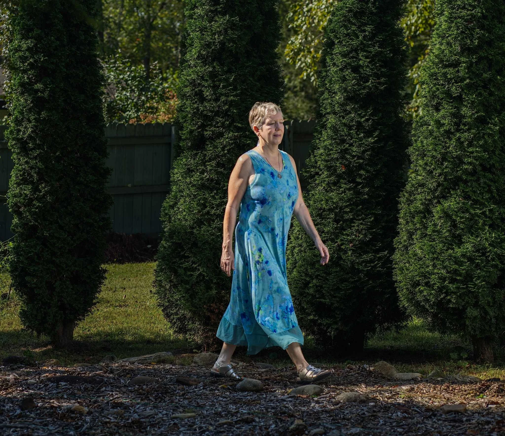 Lisa Garrett wearing a blue tie-dye dress and sandals walking outdoors in front of tall, dense evergreen trees.