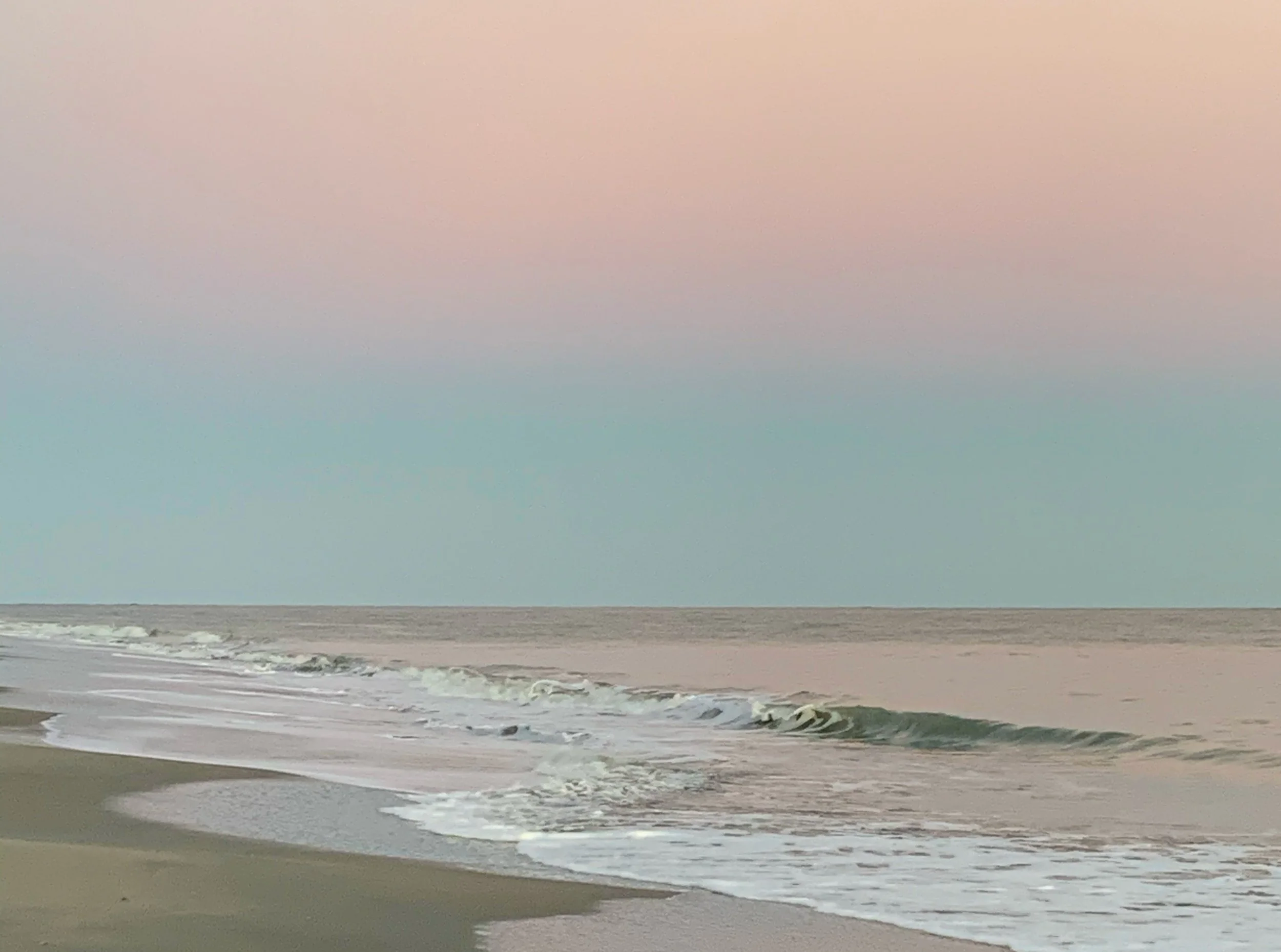 A sandy beach with small waves crashing onto the shore under a delicate pink and blue overcast sky.