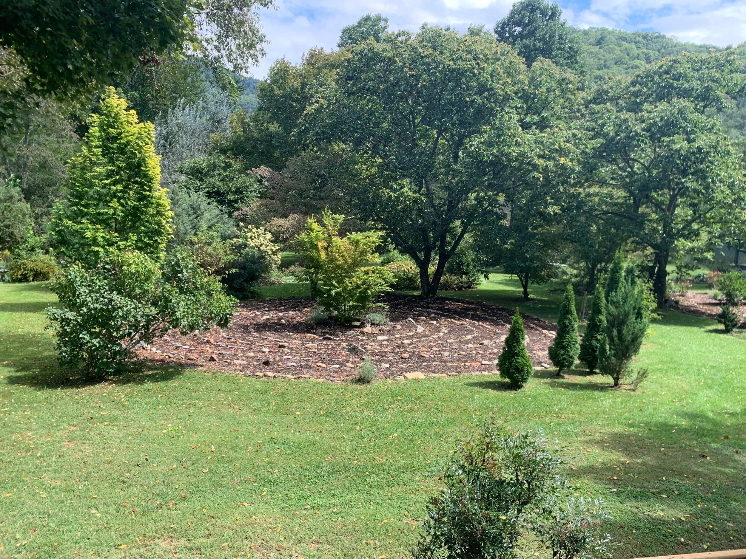 A lush, green garden with various trees, bushes, and a grassy lawn, under a partly cloudy sky.