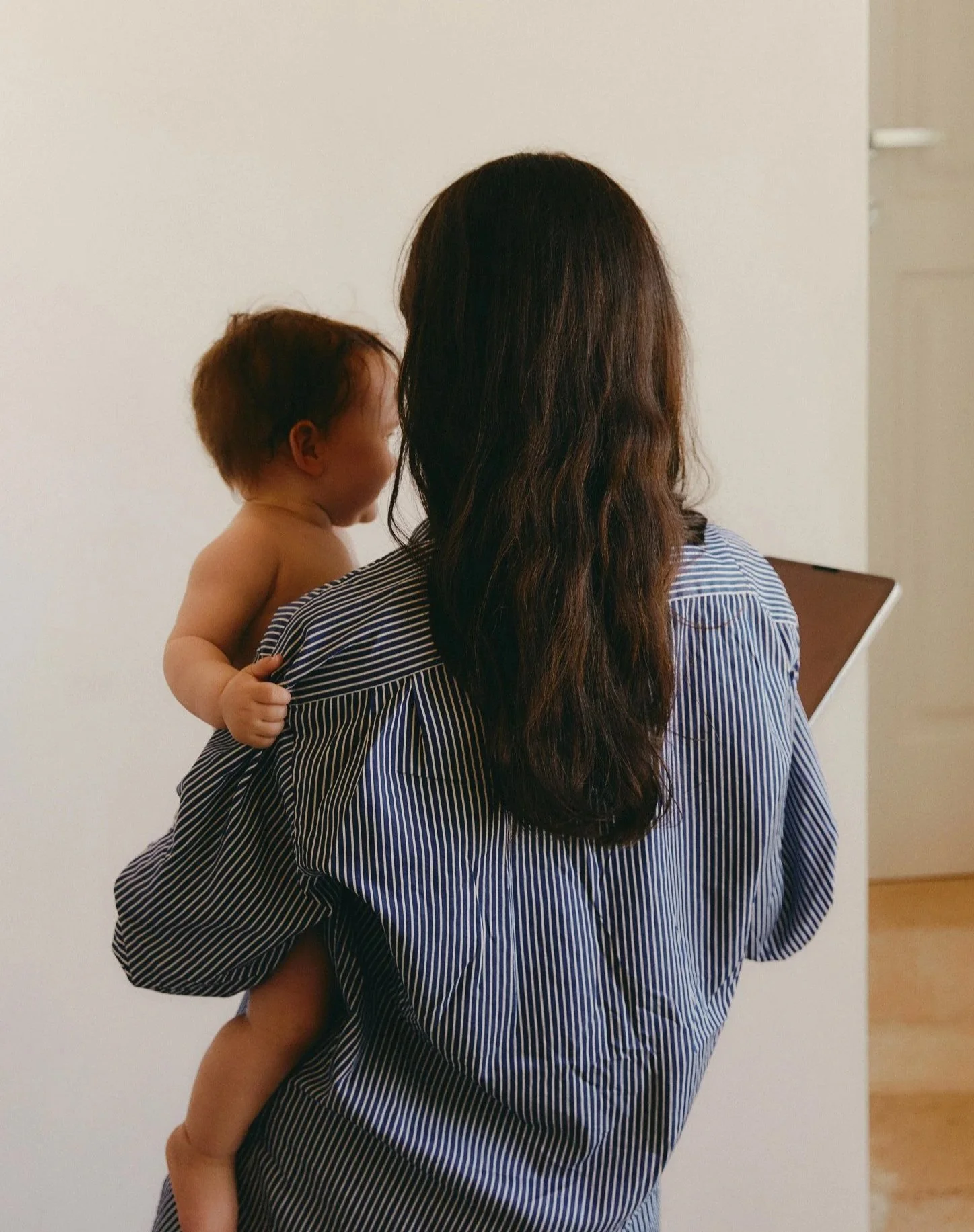 A woman with long brown hair holding a young child with brown hair and no shirt, standing against a plain white wall.