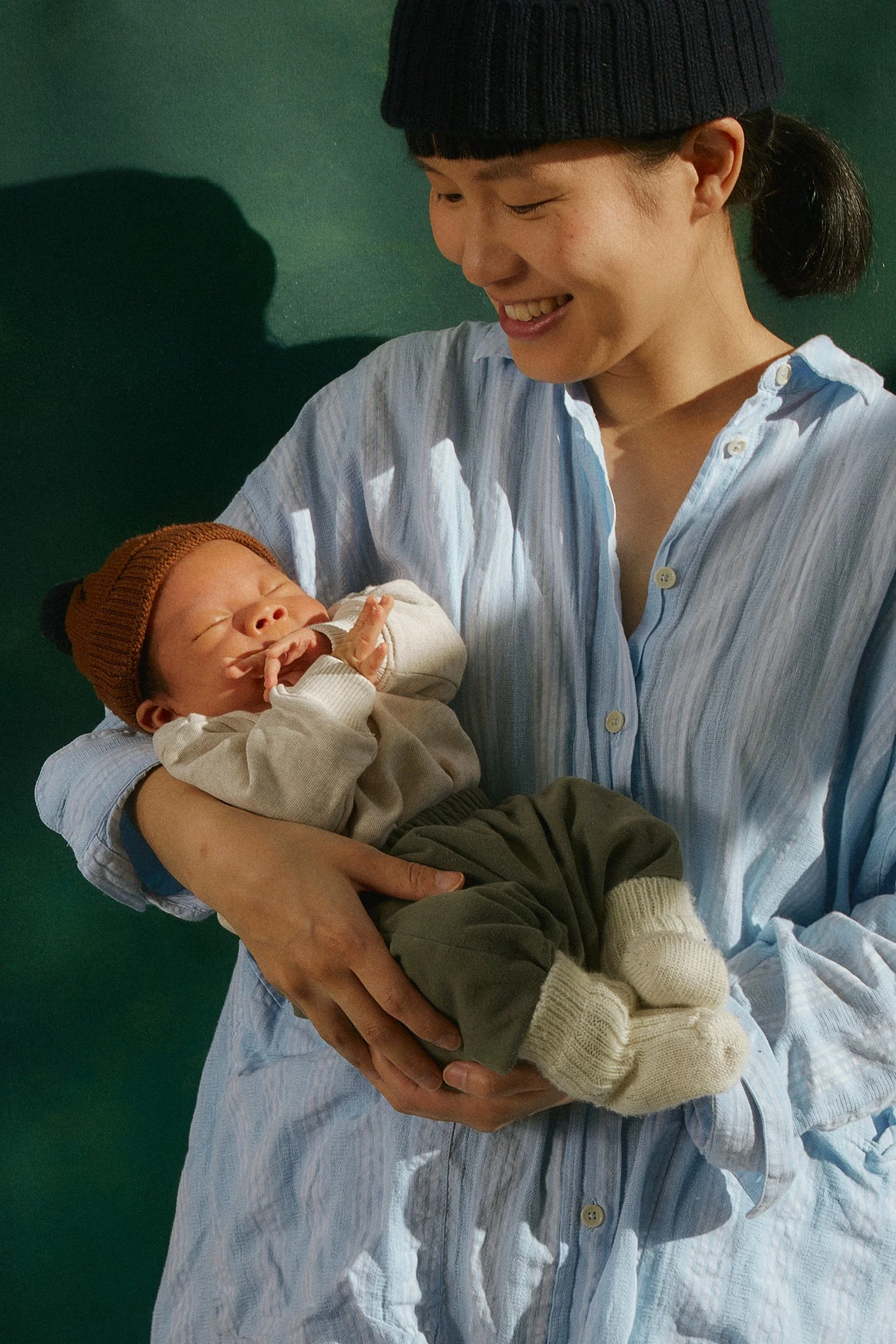 A woman with black hair in a ponytail, wearing a black knit beanie and a light blue button-up shirt, is holding a sleeping newborn baby dressed in a beige sweater, gray pants, and cream-colored knit socks, against a dark green background.