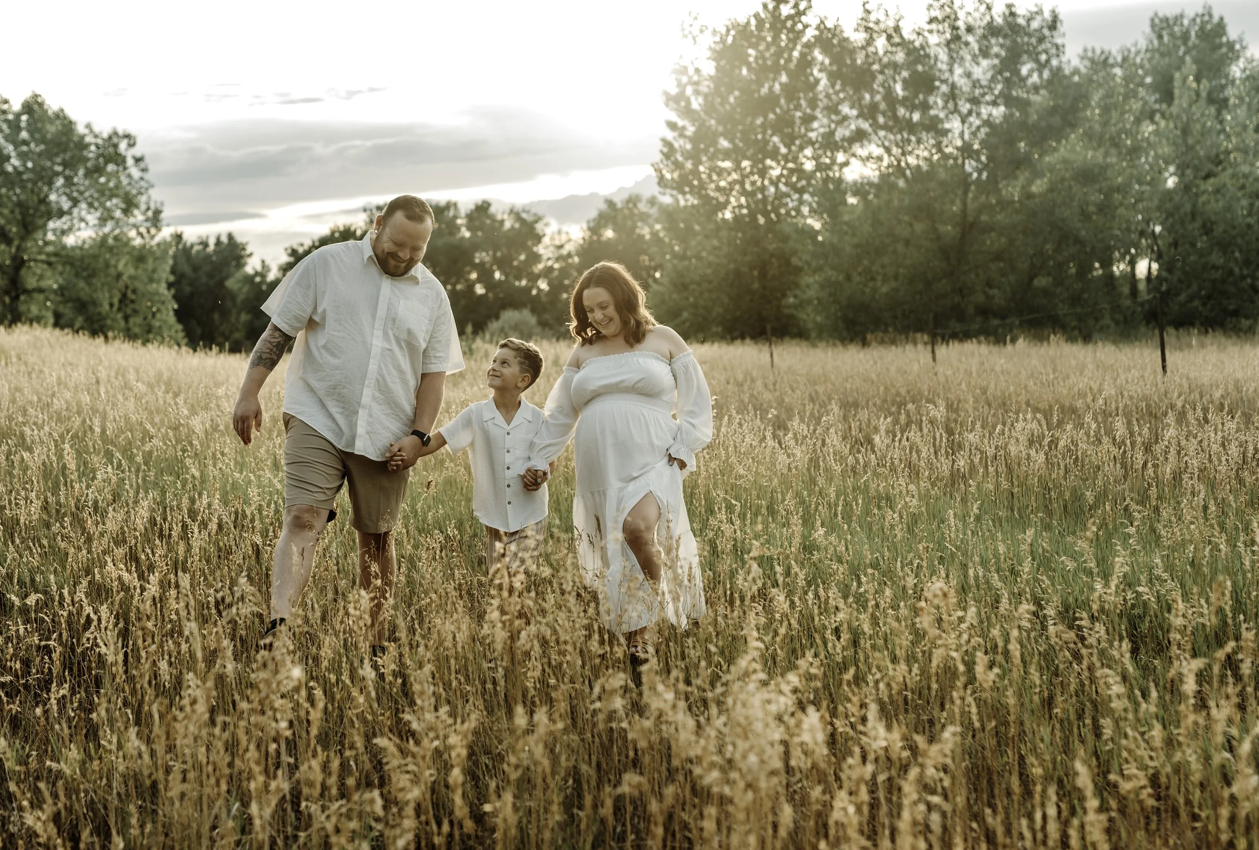 A family of three walking through a field of tall, golden grass during sunset, holding hands and smiling.
