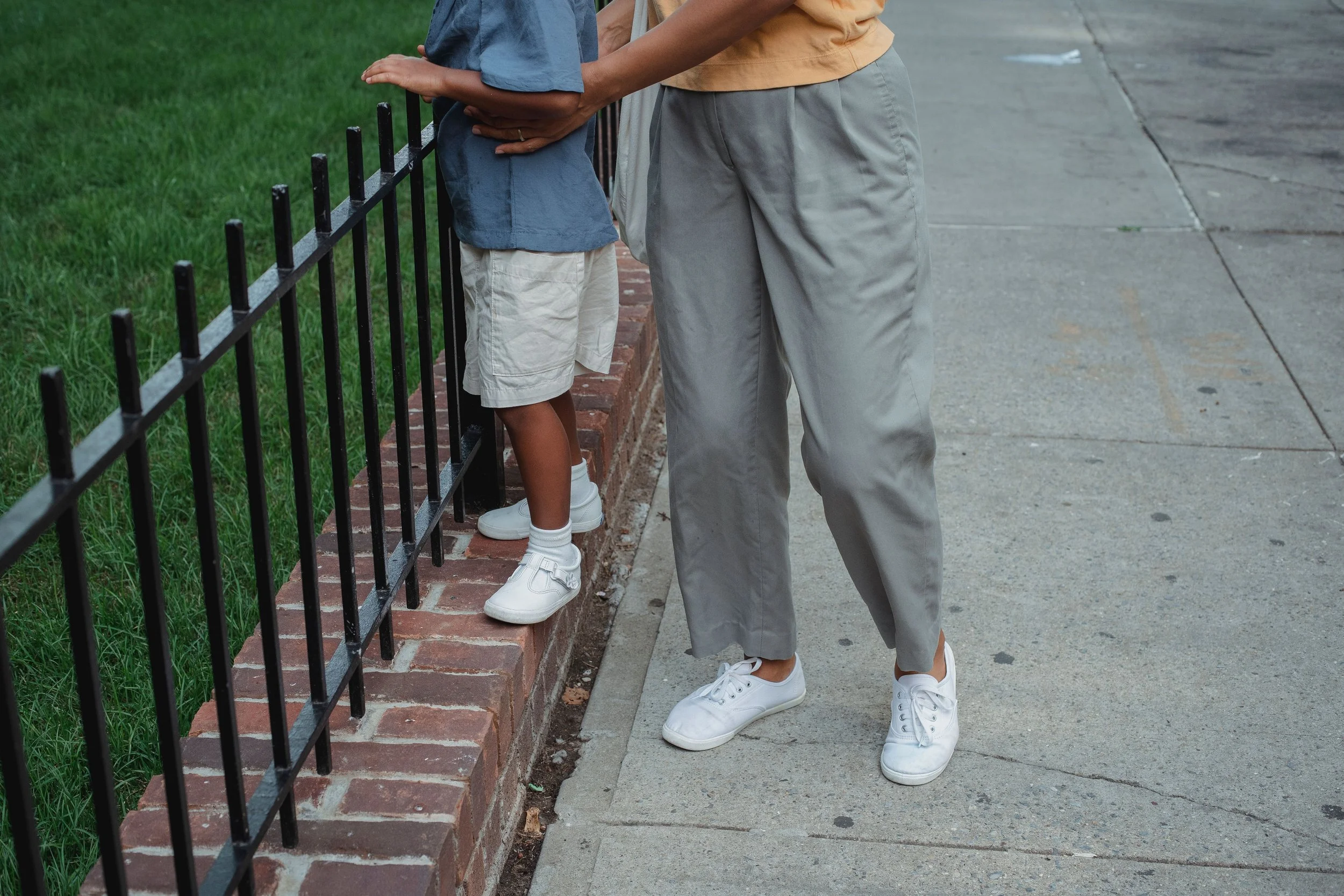 Two people, an adult and a child, standing side by side at a sidewalk, holding each other's hands on a black metal fence. The child wears a blue shirt, beige shorts, and white sneakers. The adult wears a beige shirt, light gray pants, and white sneakers.