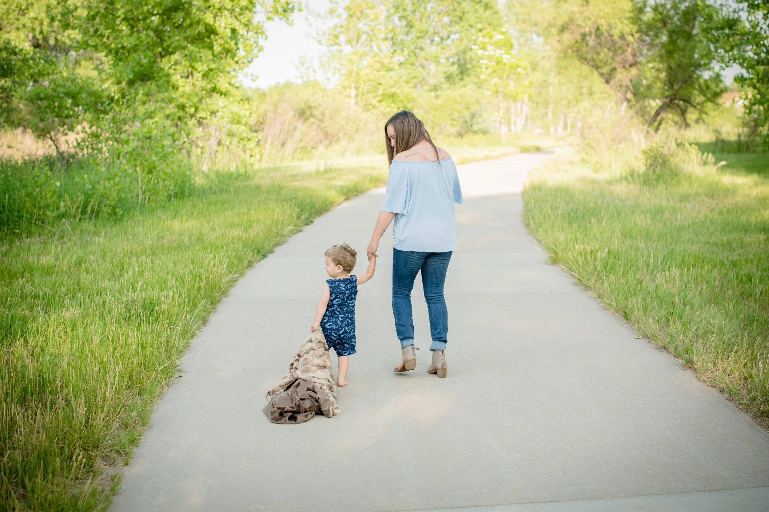 A woman and two young children walking on a paved path in a lush green park during daytime, with trees and grass on either side.