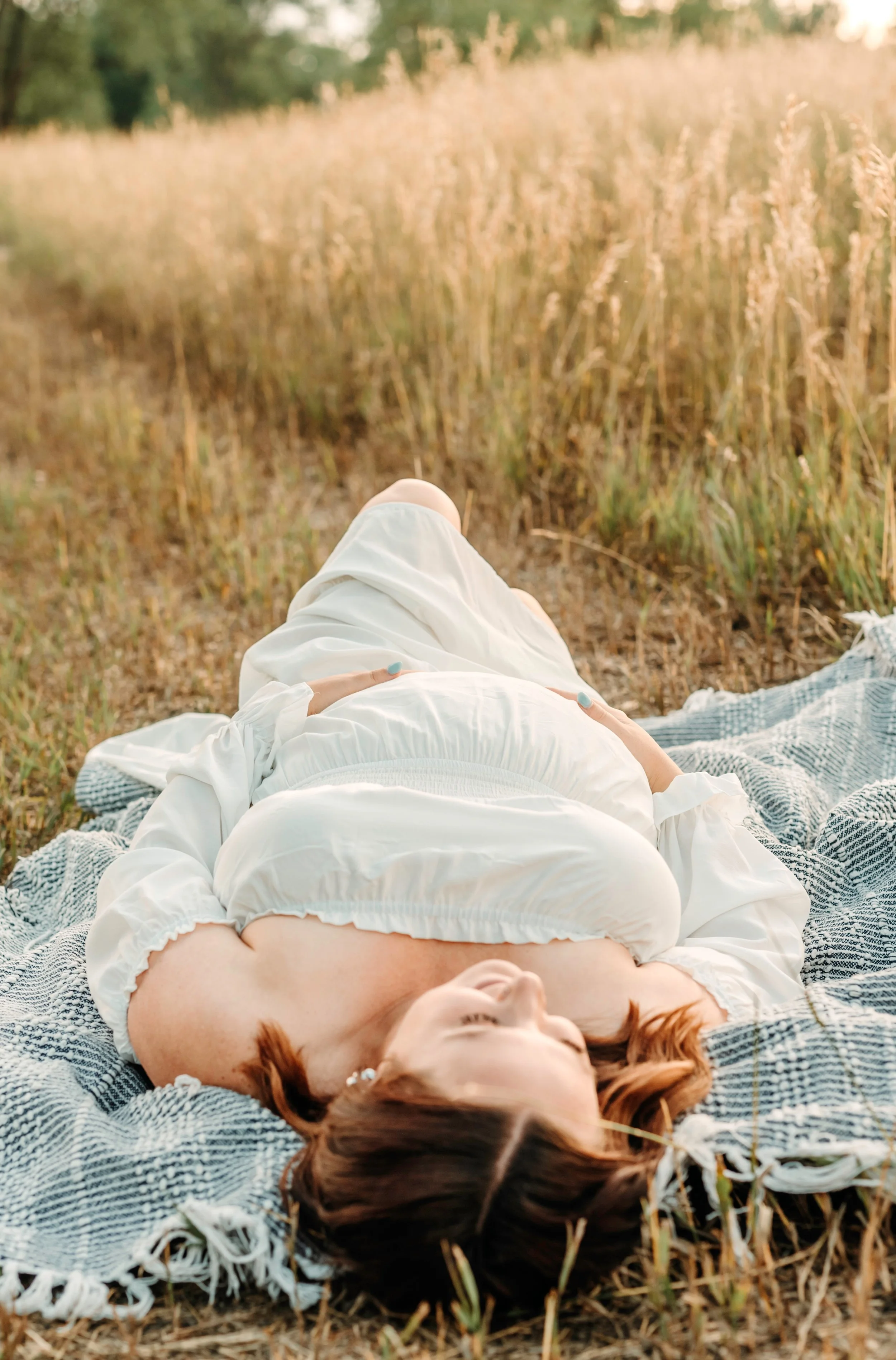 A woman with reddish-brown hair lying on her back on a checkered blanket in a grassy field with tall, golden grass, during sunset.