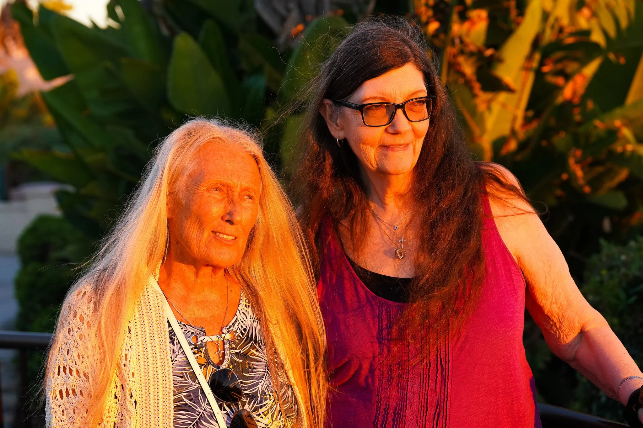 Two women with long hair sitting outdoors during sunset, smiling and looking at each other, surrounded by green plants and orange flowers.