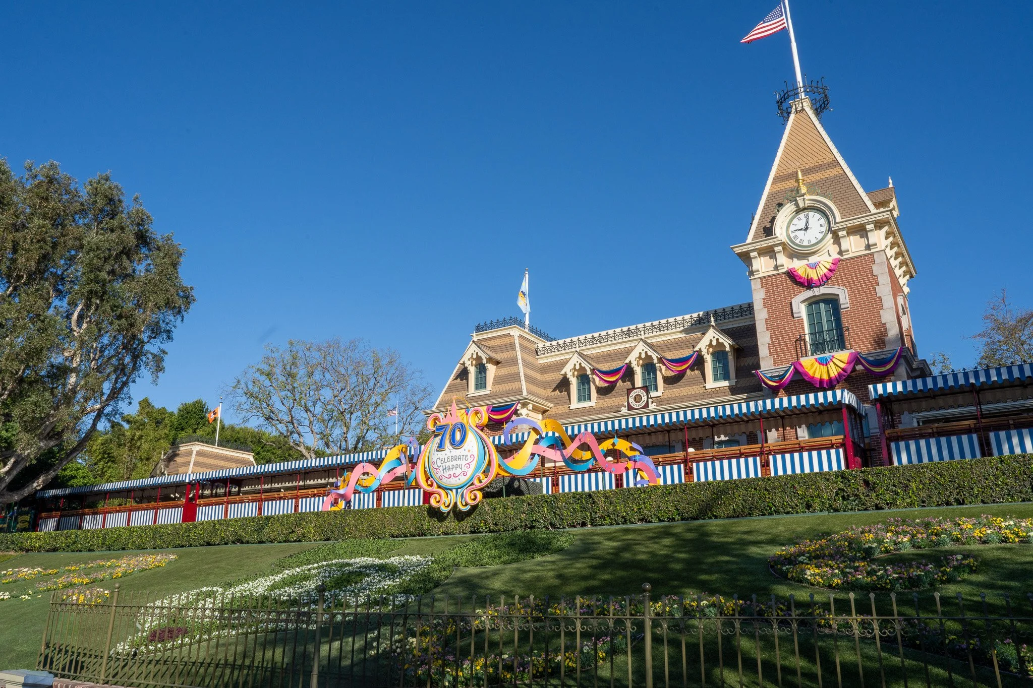 Disneyland Main Street Train Station, decorated with colorful banners celebrating 70 years, with a clock tower, an American flag, a blue sky, and trees in the background.