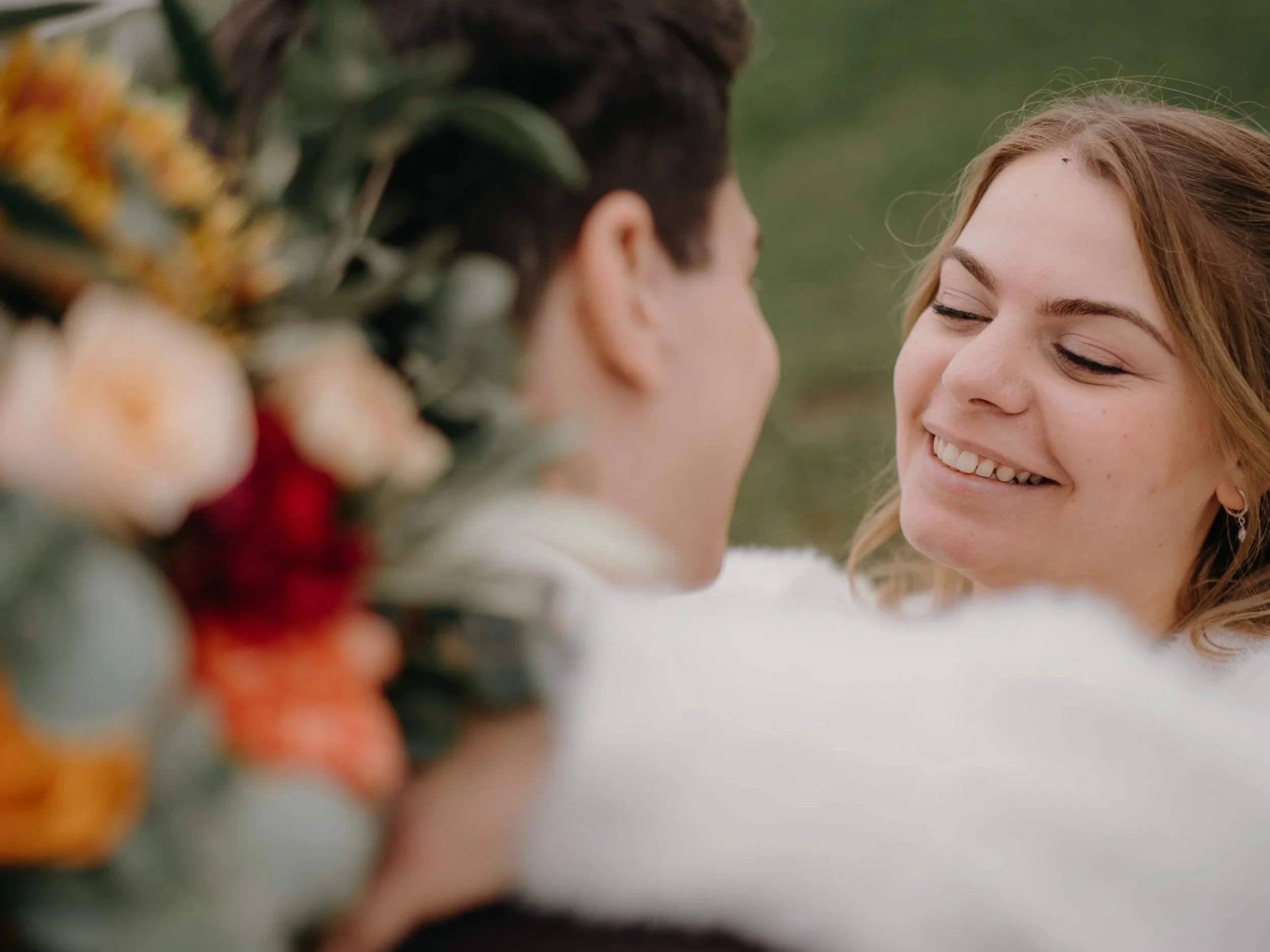 Ein lächelndes Mädchen mit roten Haaren und einem weißen Kleid, das einem Mann gegenübersitzt. Im Vordergrund befindet sich eine bunte Blumen-Duftkränze, die unscharf sind.