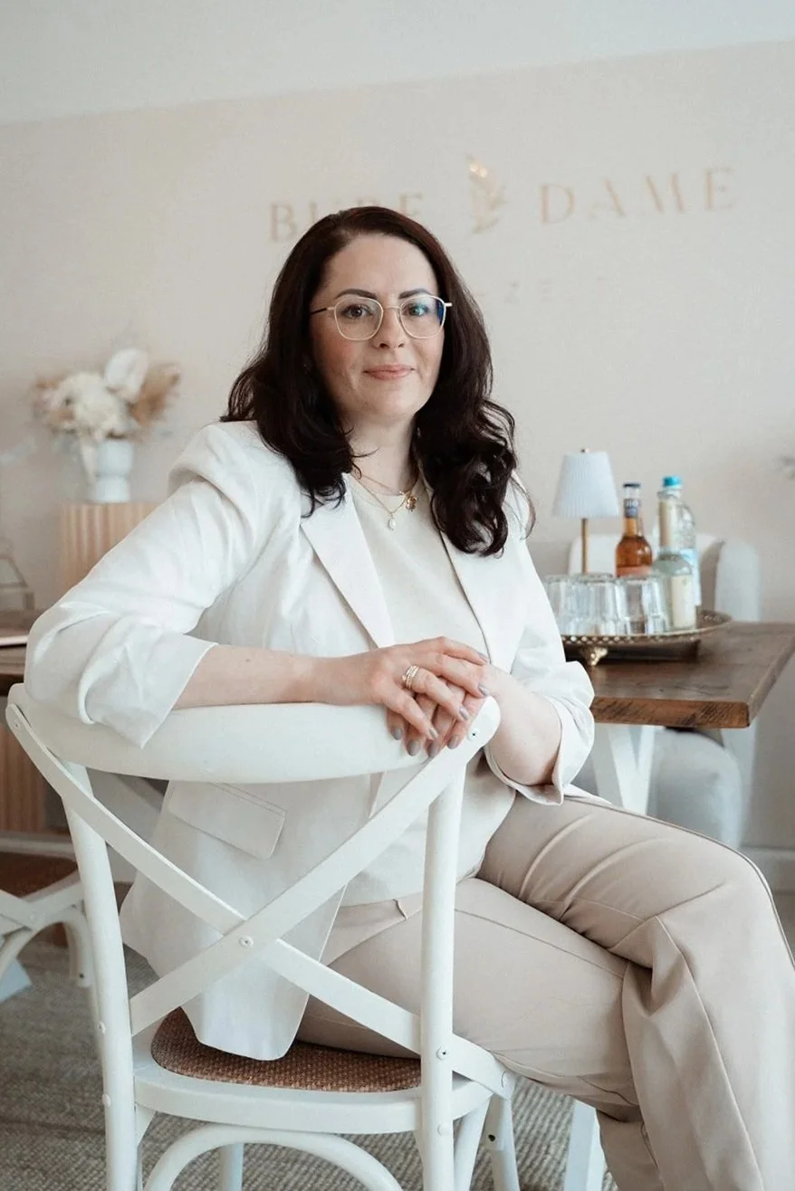 Eine Frau mit Brille sitzt auf einem weißen Stuhl in einem modernen, hellen Raum, hinter ihr ist eine Bar mit Flaschen und Gläsern, im Hintergrund eine Vase mit Blumen und eine Lampe.