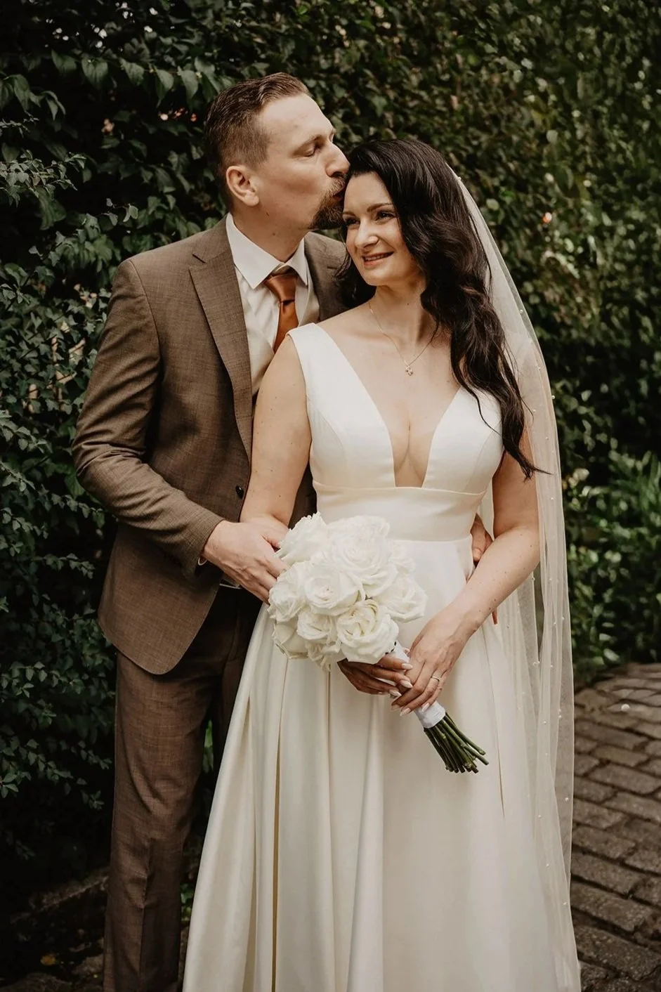 Ein Brautpaar bei der Hochzeit: Der Bräutigam in Anzug küsst die Braut, die ein weißes Brautkleid trägt und einen Blumenstrauß hält, vor einer grünen Hecke.
