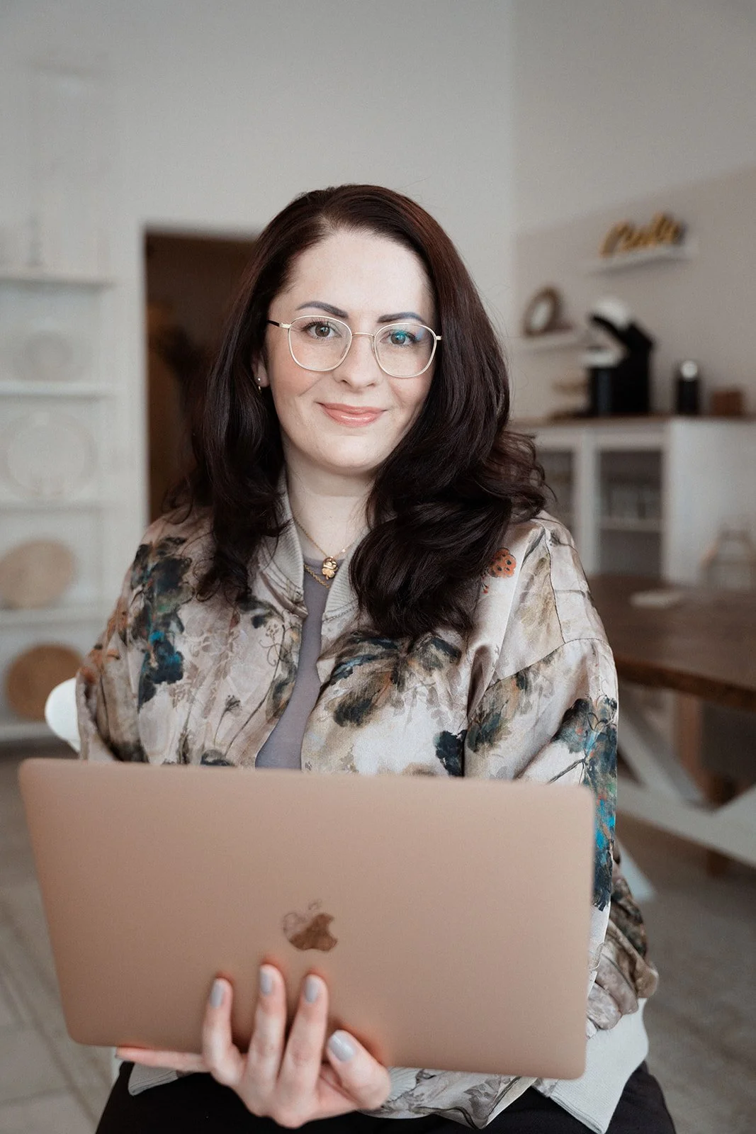 Frau mit Brille und dunklem Haar, die einen Laptop hält, im Hintergrund eine Küche.