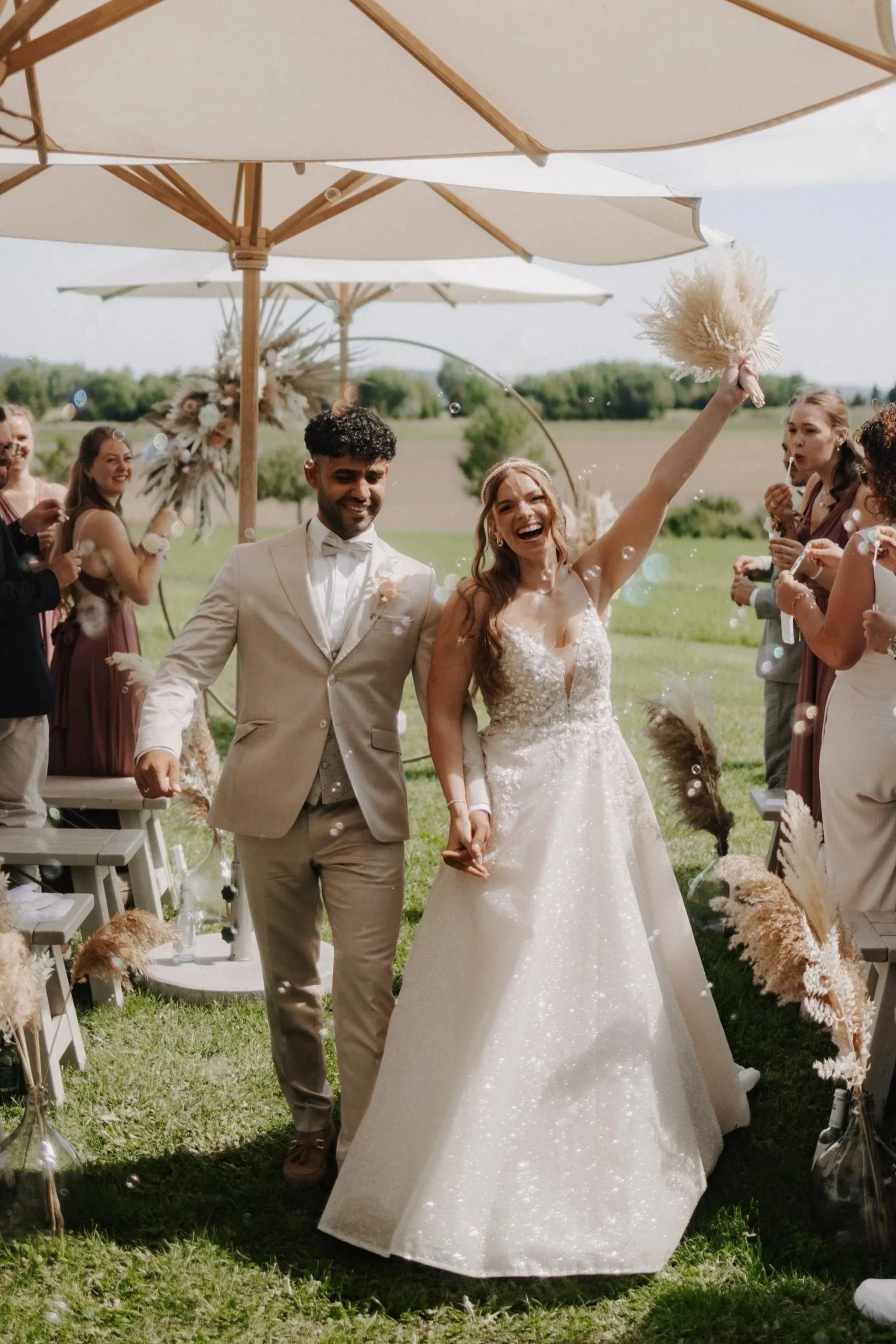 Ein frisch verheiratetes Paar läuft Händchen haltend durch eine Hochzeitsparty im Freien, umgeben von Freunden und Familie, mit Sonnenschirmen im Hintergrund.