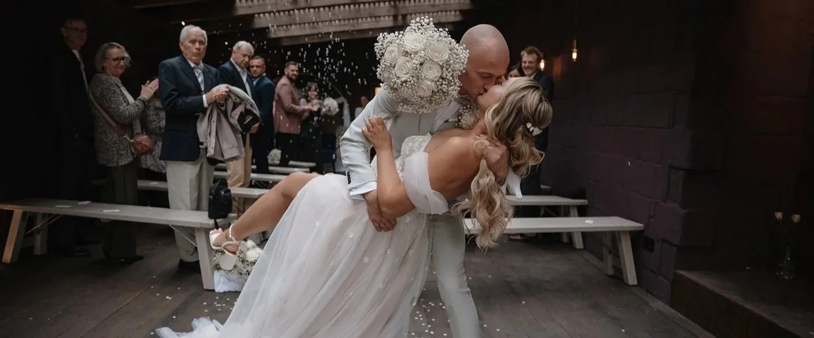 Braut und Bräutigam küssen sich bei ihrer Hochzeit im Dunkeln, während Gäste im Hintergrund zusehen.