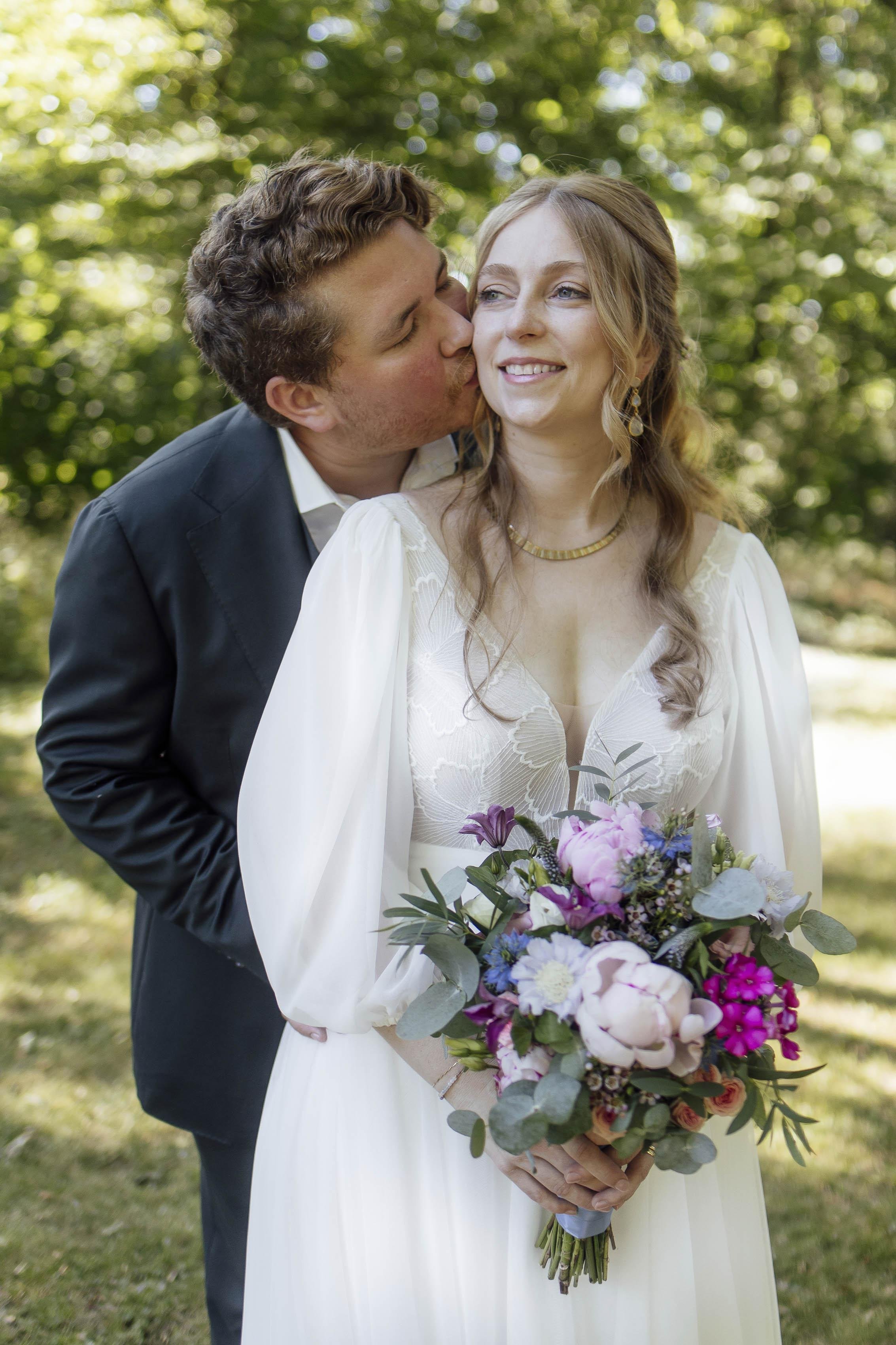 Ein Mann küsst eine lächelnde Frau bei einer Hochzeit im Freien, sie hält einen bunten Blumenstrauß.
