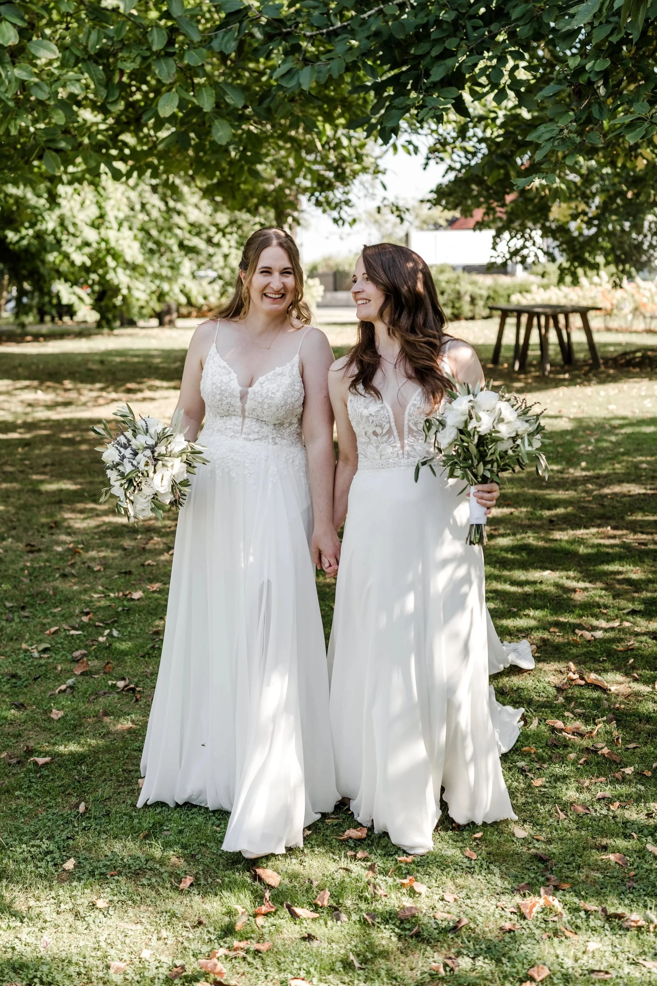 Zwei Frauen in Hochzeitskleidern stehen auf einer Wiese unter einem Baum, halten Hände und lachen. Sie tragen weiße Brautkleider und halten Blumensträuße.