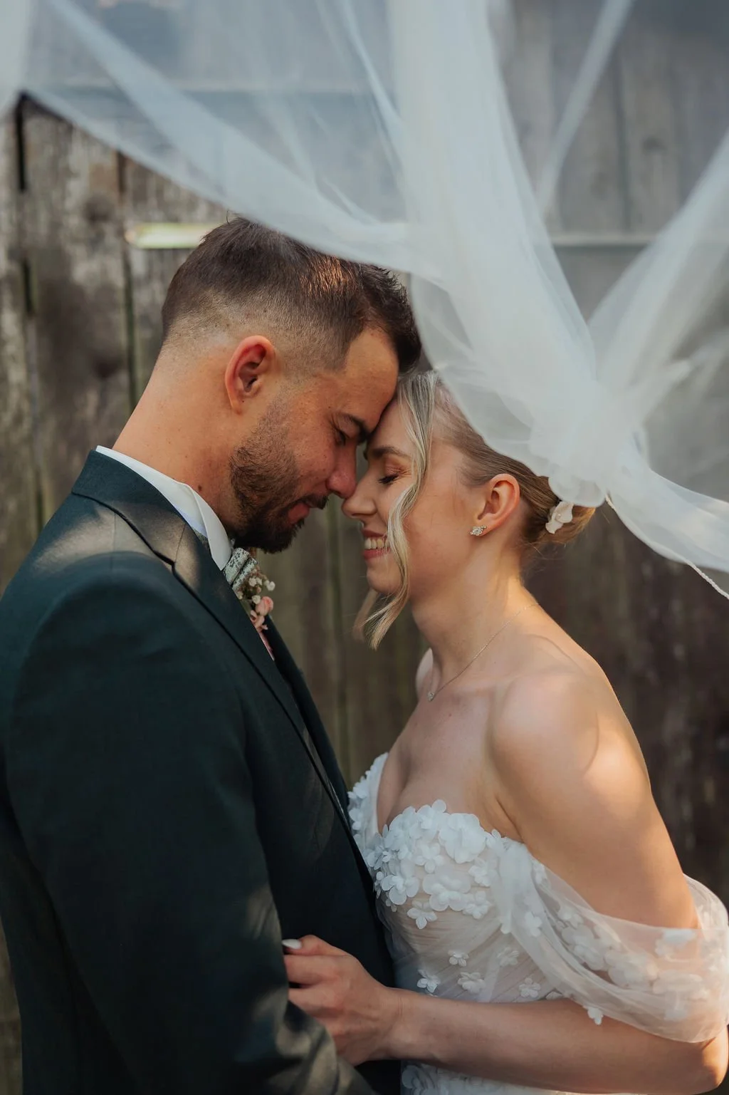 Ein verlobtes Paar bei einer Hochzeit, eng aneinandergeschmiegt, unter einem weißen Schleier, lächelnd, vor einer Holzwand.