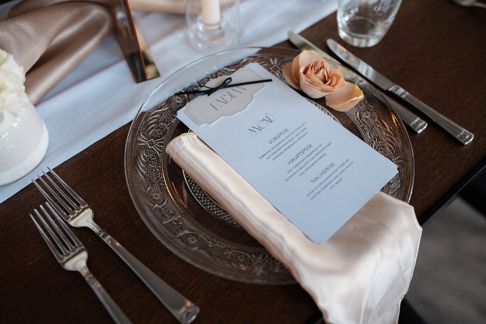 Tisch setting mit einer Menükarte, einem weißen Stoffserviette, einem Rosenblütenblatt, Besteck und Trinkglas auf einem dunklen Holztisch, gedeckter Tisch für ein festliches Essen.