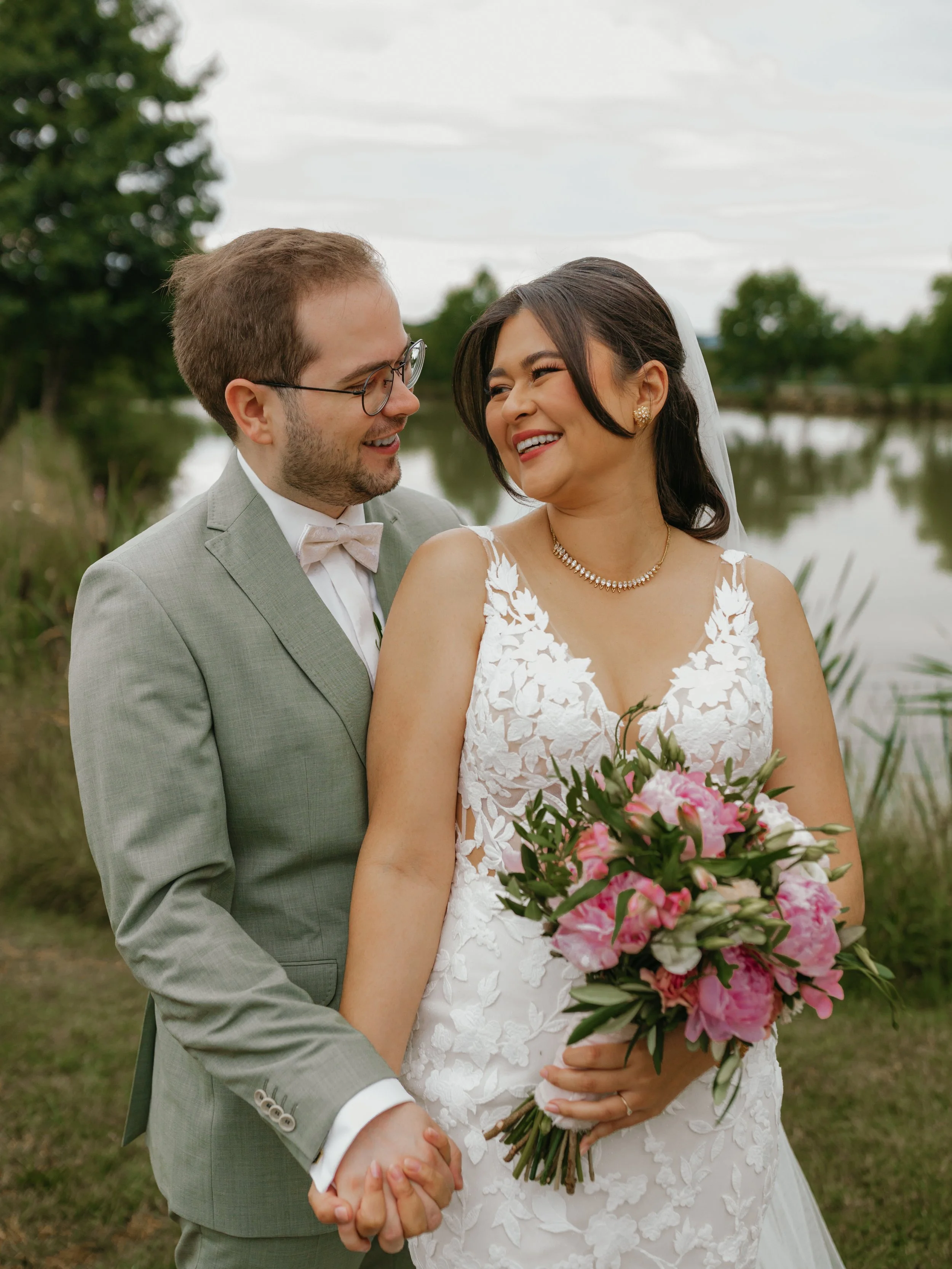 Ein Brautpaar beim Spaziergang am Fluss, lachend und Händchen haltend. Die Braut trägt ein weißes Kleid mit Blumenmuster und hält einen Blumenstrauß. Der Bräutigam trägt einen hellgrauen Anzug mit Fliege.