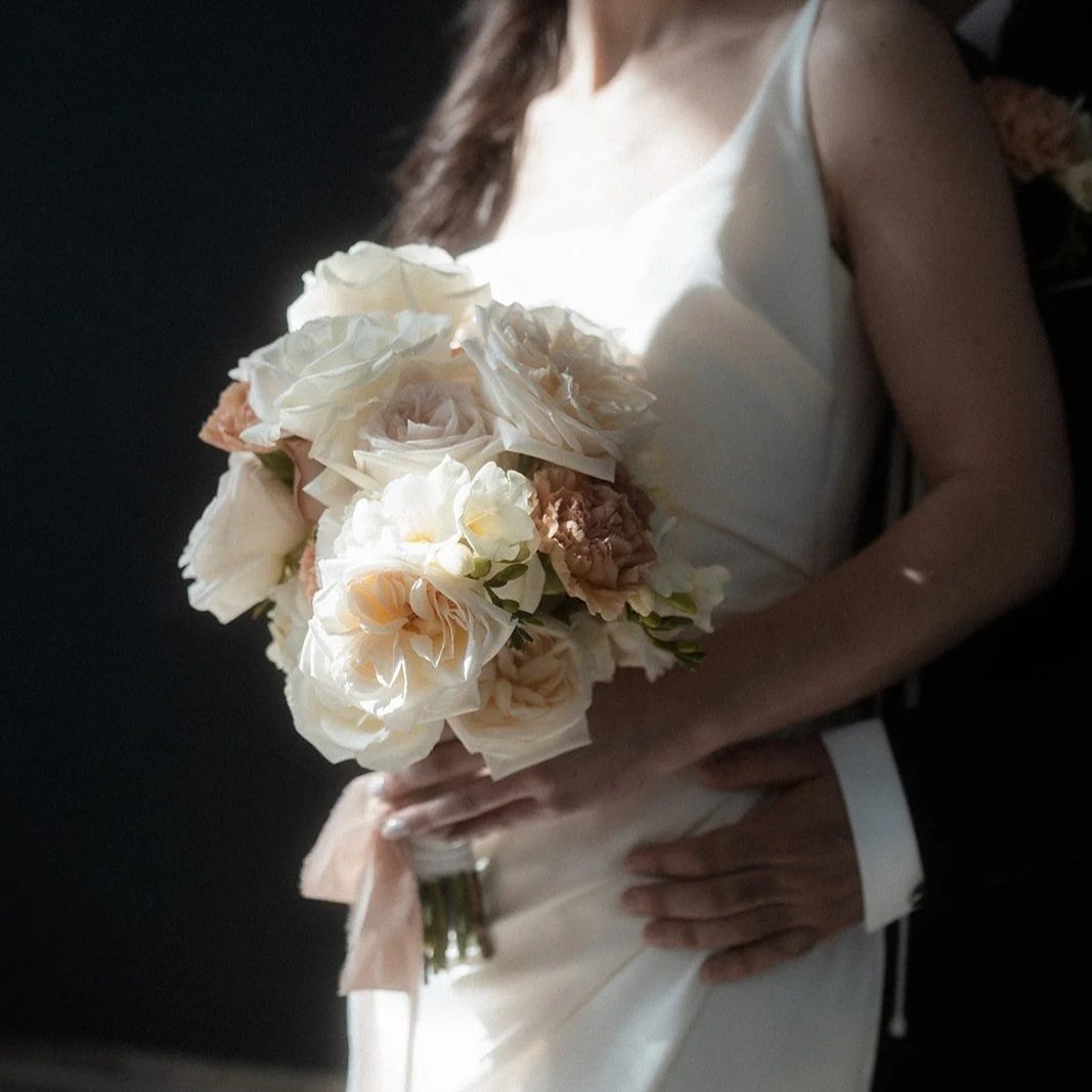 Frau in einem weißen Kleid hält einen Brautstrauß mit weißen und rosafarbenen Blumen vor einem dunklen Hintergrund.