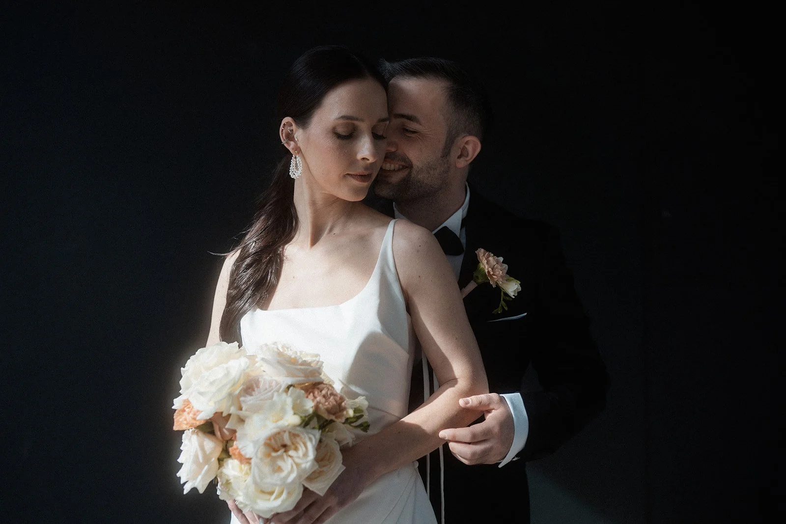 Ein frisch vermähltes Paar bei ihrer Hochzeit, die Braut hält einen Blumenstrauß, sie trägt ein weißes Kleid, der Bräutigam trägt einen schwarzen Anzug mit Fliege, steht vor dunklem Hintergrund.