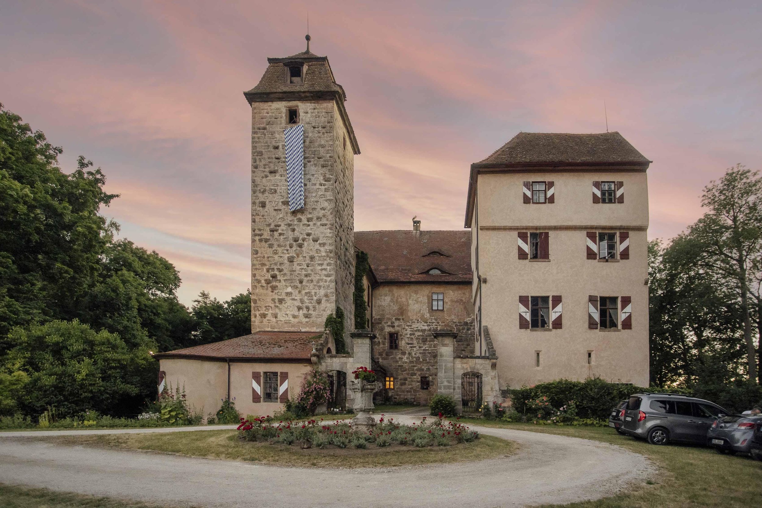 Historisches Gebäude mit einem hohen Turm, bunte Fensterläden und umgebende Natur bei Sonnenuntergang.