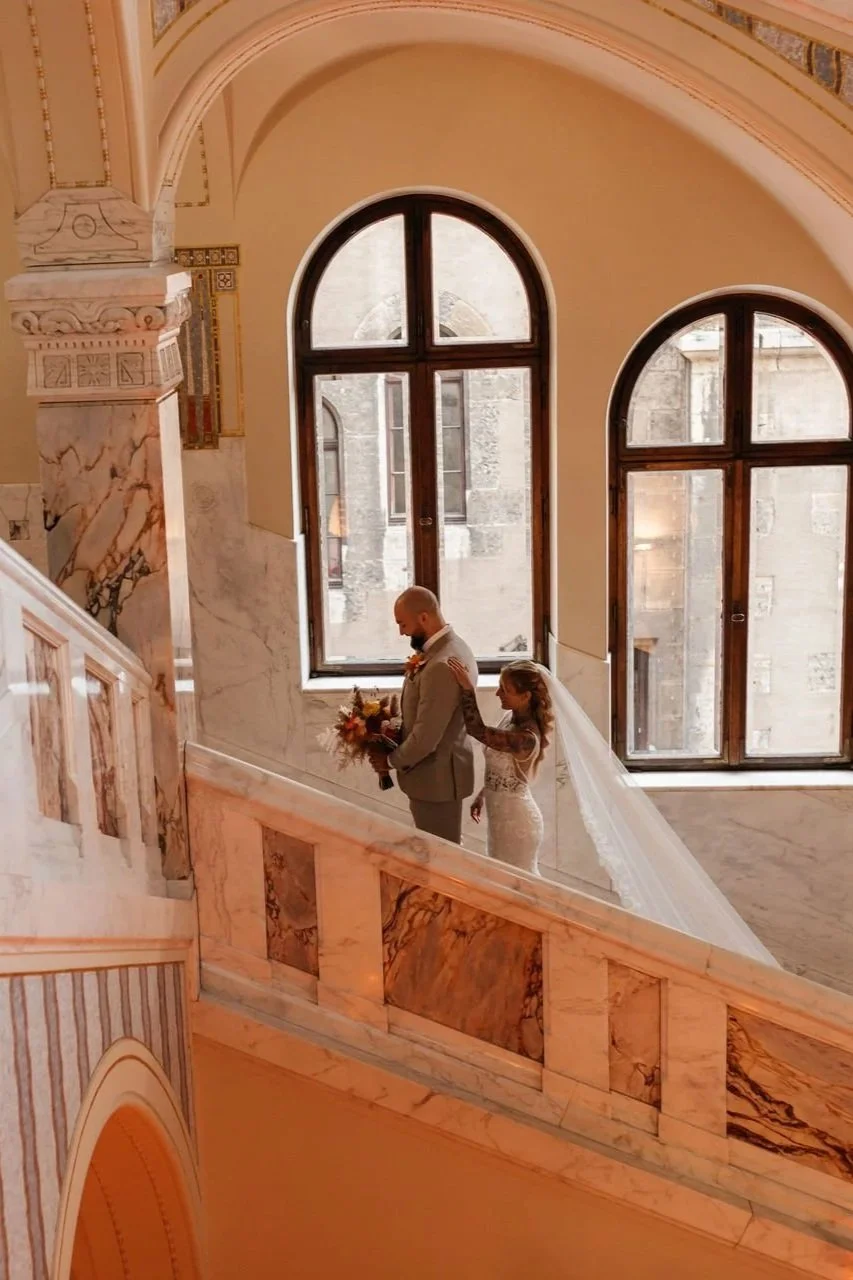 Braut und Bräutigam auf einer Marmortreppe in einer eleganten, historischen Gebäudehalle, mit großen arched Fenstern im Hintergrund.