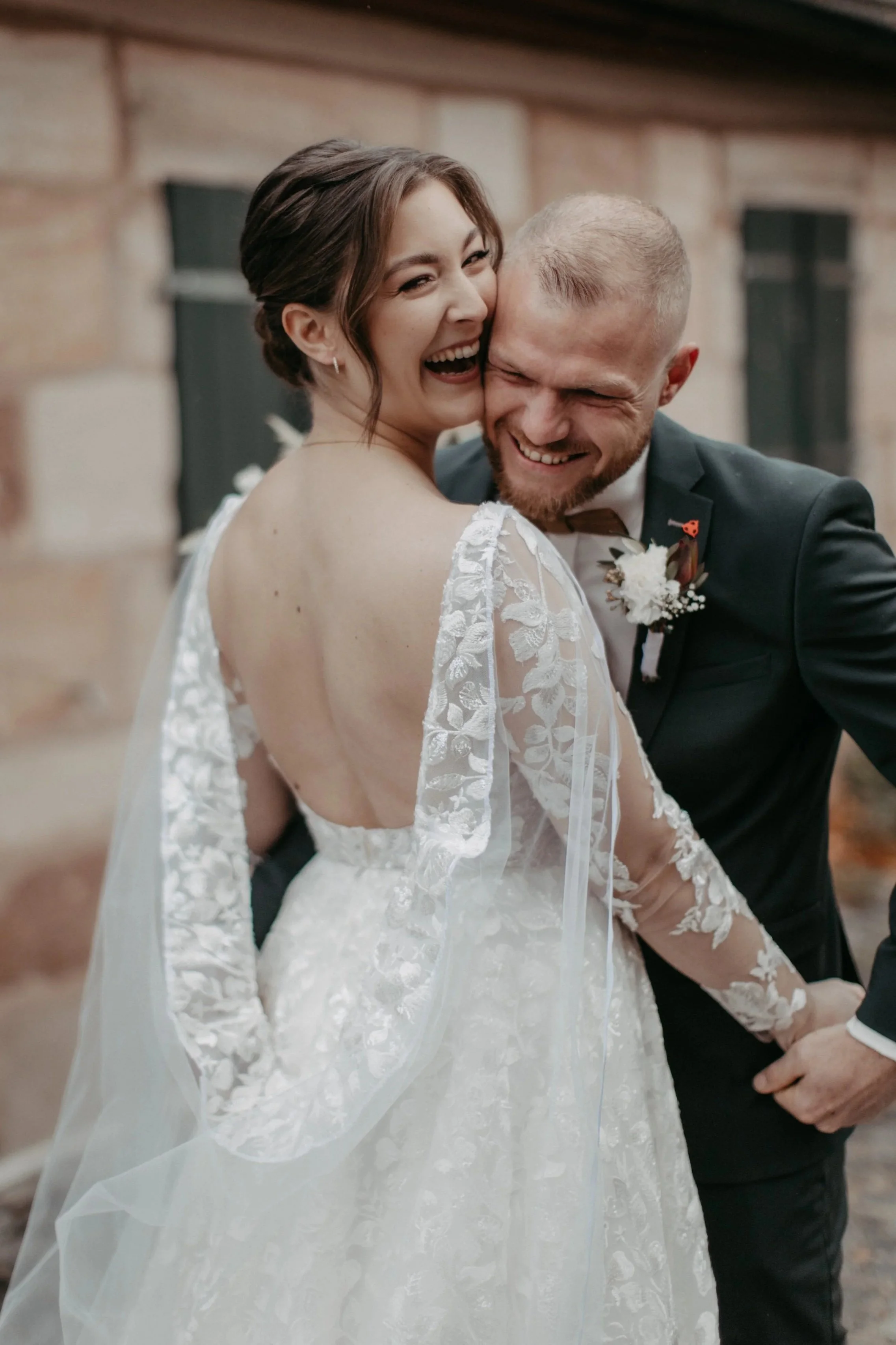 Das Bild zeigt ein glückliches Hochzeitspaar, bei dem die Braut in einem weißen Brautkleid lacht und sich an den Bräutigam schmiegt, der einen dunklen Anzug trägt, in einer rustikalen Umgebung mit Backsteinmauer im Hintergrund.