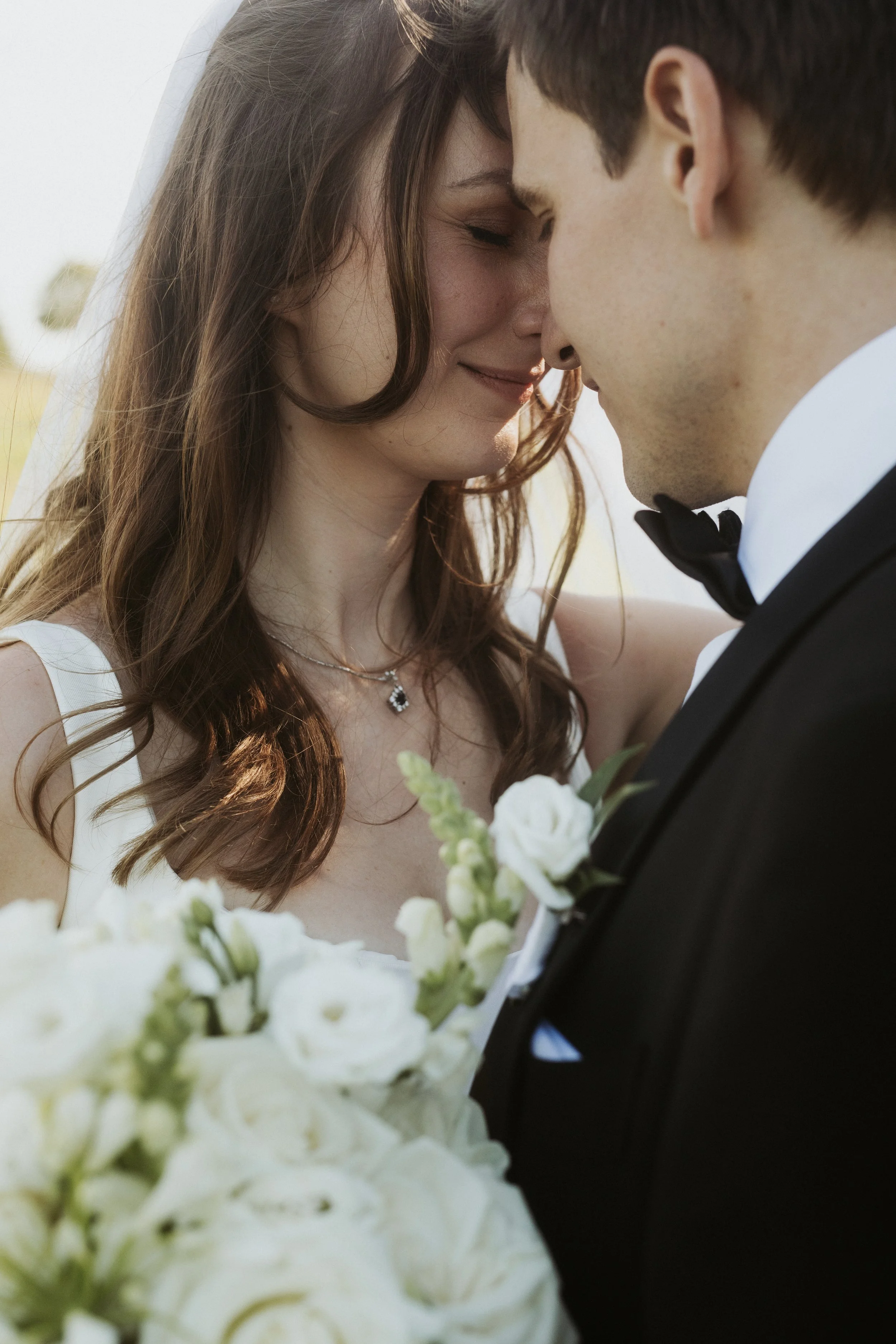 Braut und Bräutigam stehen sich mit Stirn an Stirn, beide lächeln zärtlich, bei einer Hochzeit, im Hintergrund Hellblau und Grün.