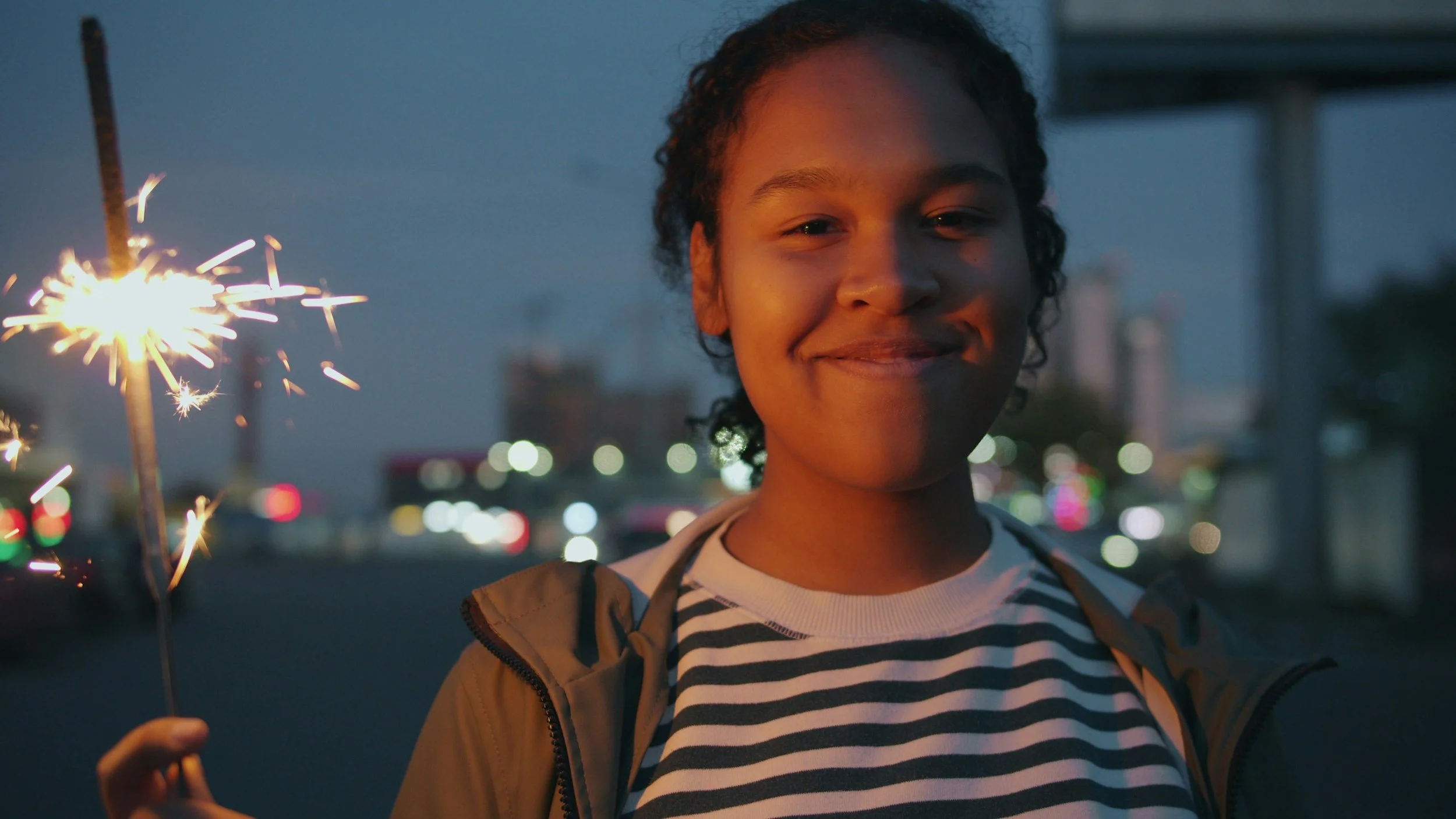 A young person smiling as they hold a lit sparkler in an outdoor setting during dusk or evening, with city lights in the background.