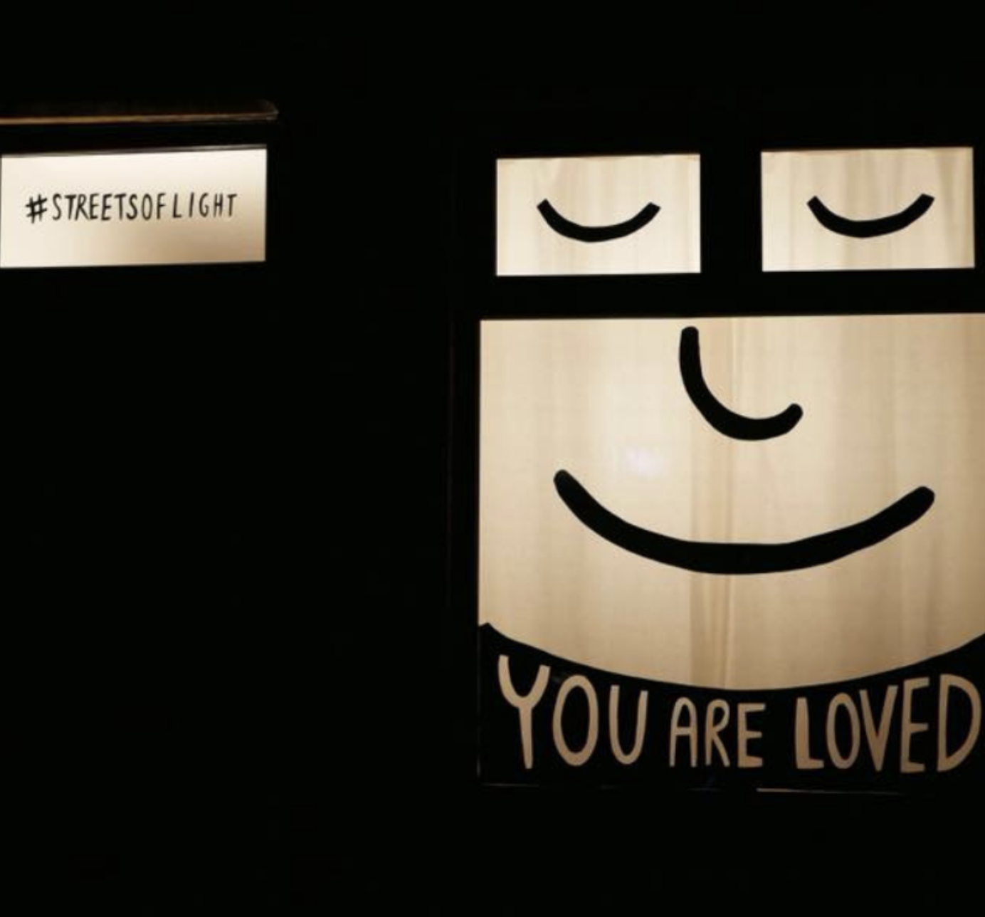 A illuminated wall sign with a smiling face and the words "YOU ARE LOVED" underneath. There is also a smaller sign that says "#STREETSOFLIGHT".