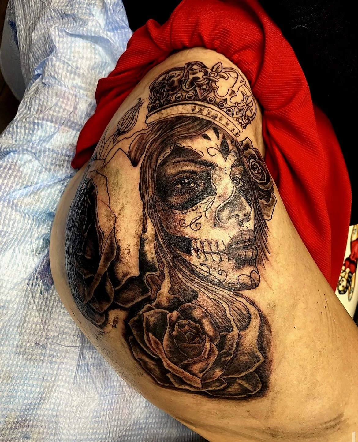 Black and grey tattoo of a woman's face with a skull makeup design, wearing a decorative crown, accompanied by roses and a hand making a gesture, on a person's thigh.