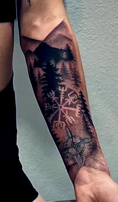 Tattoo of mountains, pine trees, and a Nordic symbol on a person's forearm.