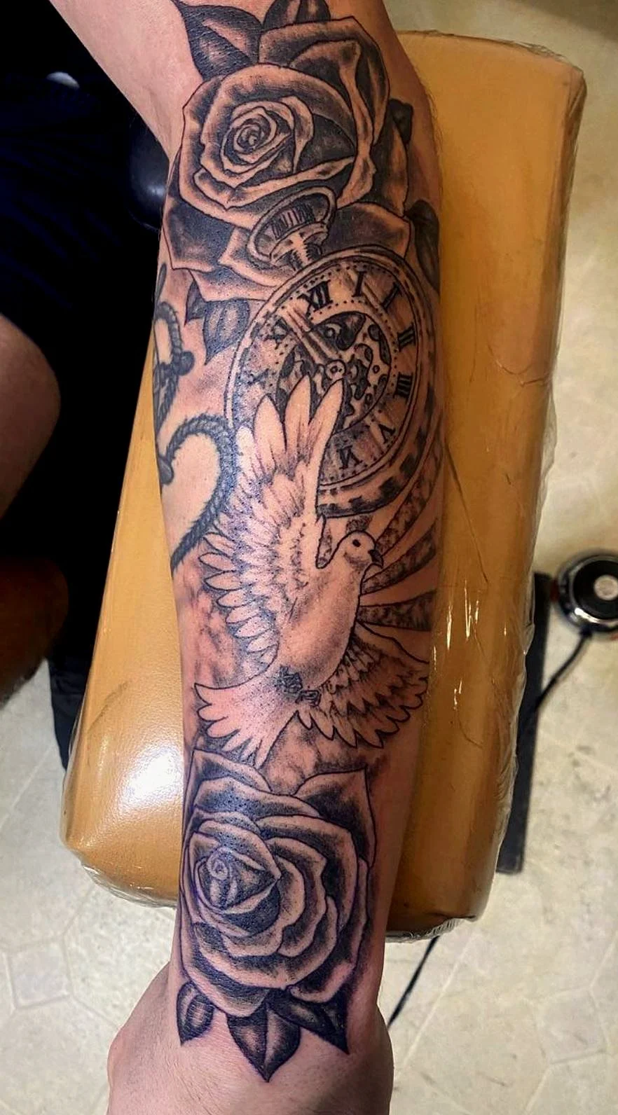 Tattoo on person's arm featuring roses, a vintage pocket watch, a dove, and a finger indicating the watch.