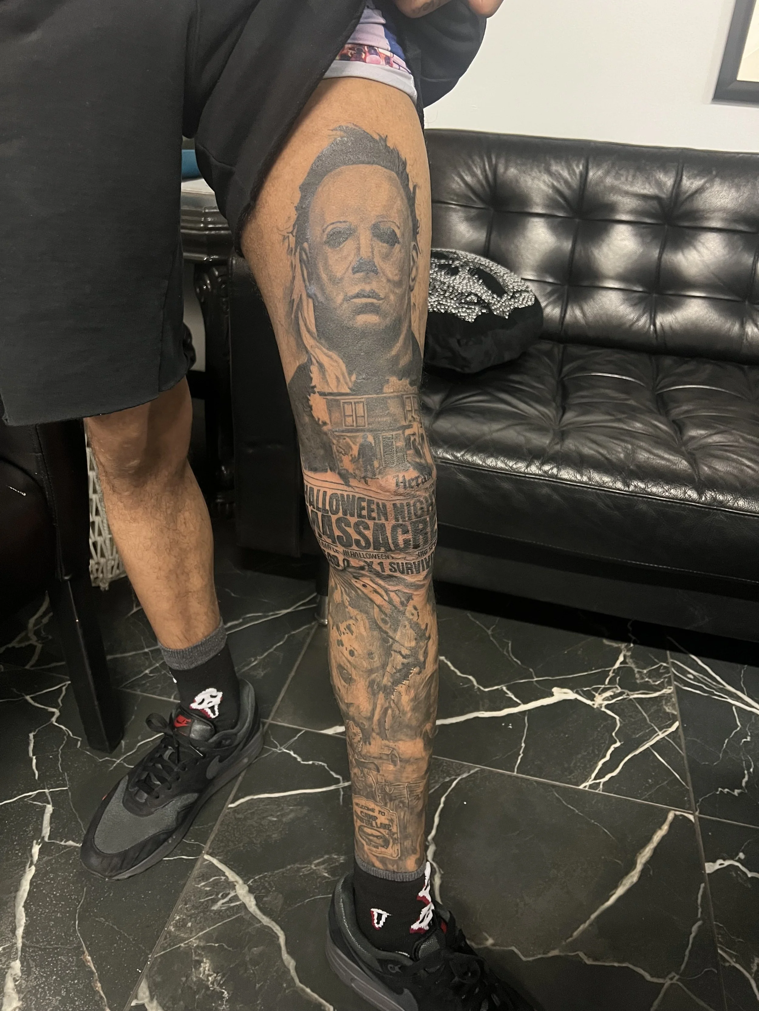 A person's leg with a detailed tattoo of a caricatured face of Michael Myers from the Halloween movie series on the thigh, and text tattoos on the lower leg including "Halloween Massacre." The person is wearing black shorts, black sneakers, and socks