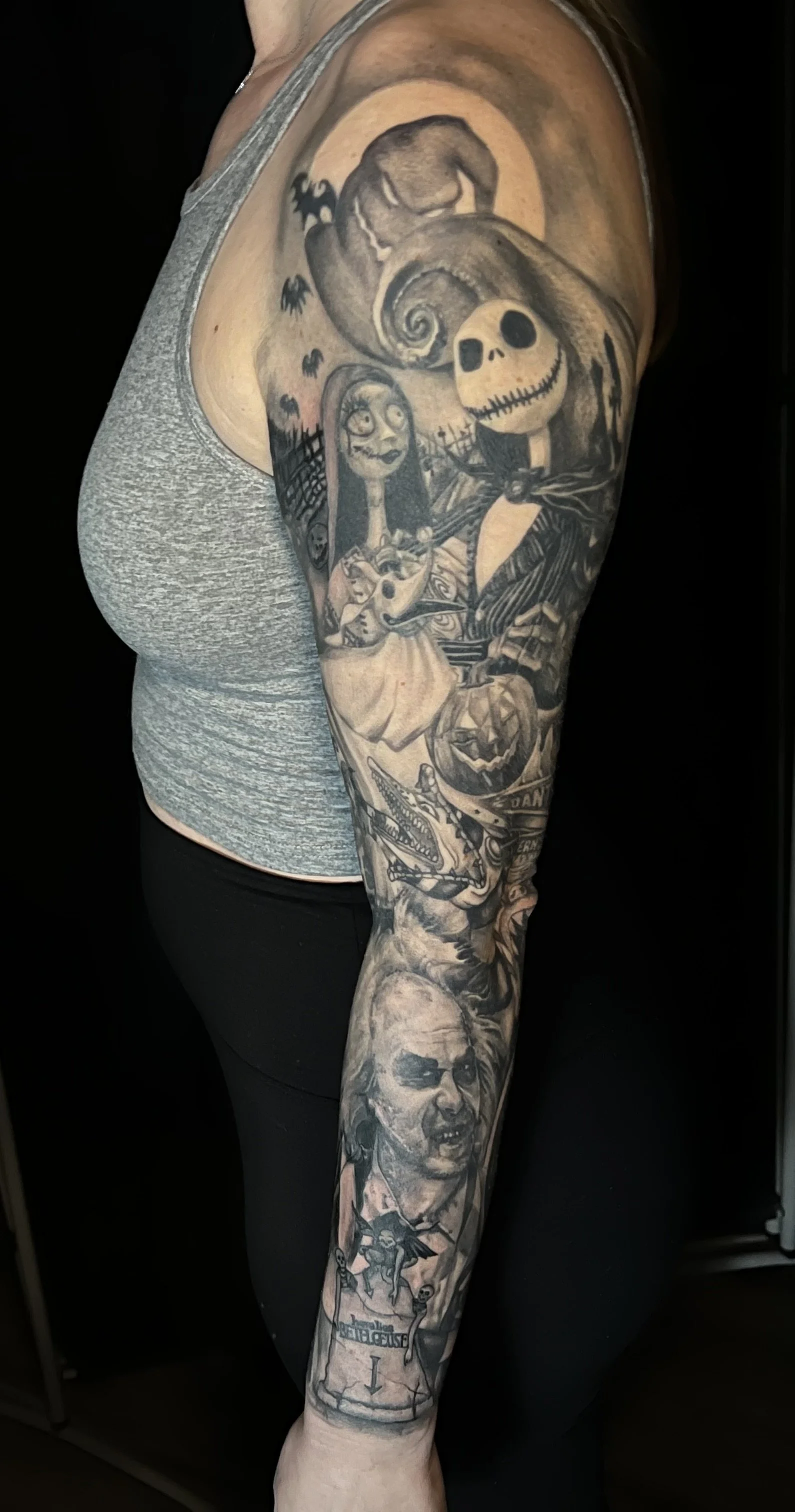 Detailed black and gray tattoo sleeve featuring cartoon characters, Halloween elements, skulls, monsters, and a clock on a person's arm.