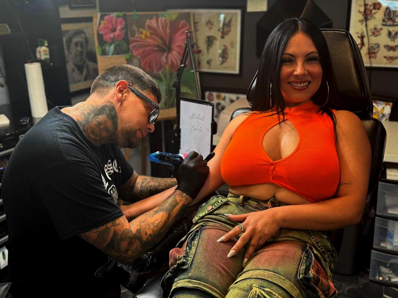 A woman with dark hair, wearing an orange sleeveless top and camouflage pants, sitting in a tattoo chair and smiling as a tattoo artist tattoos her arm. The tattoo artist, wearing glasses and black gloves, has tattoos on his arms and neck.