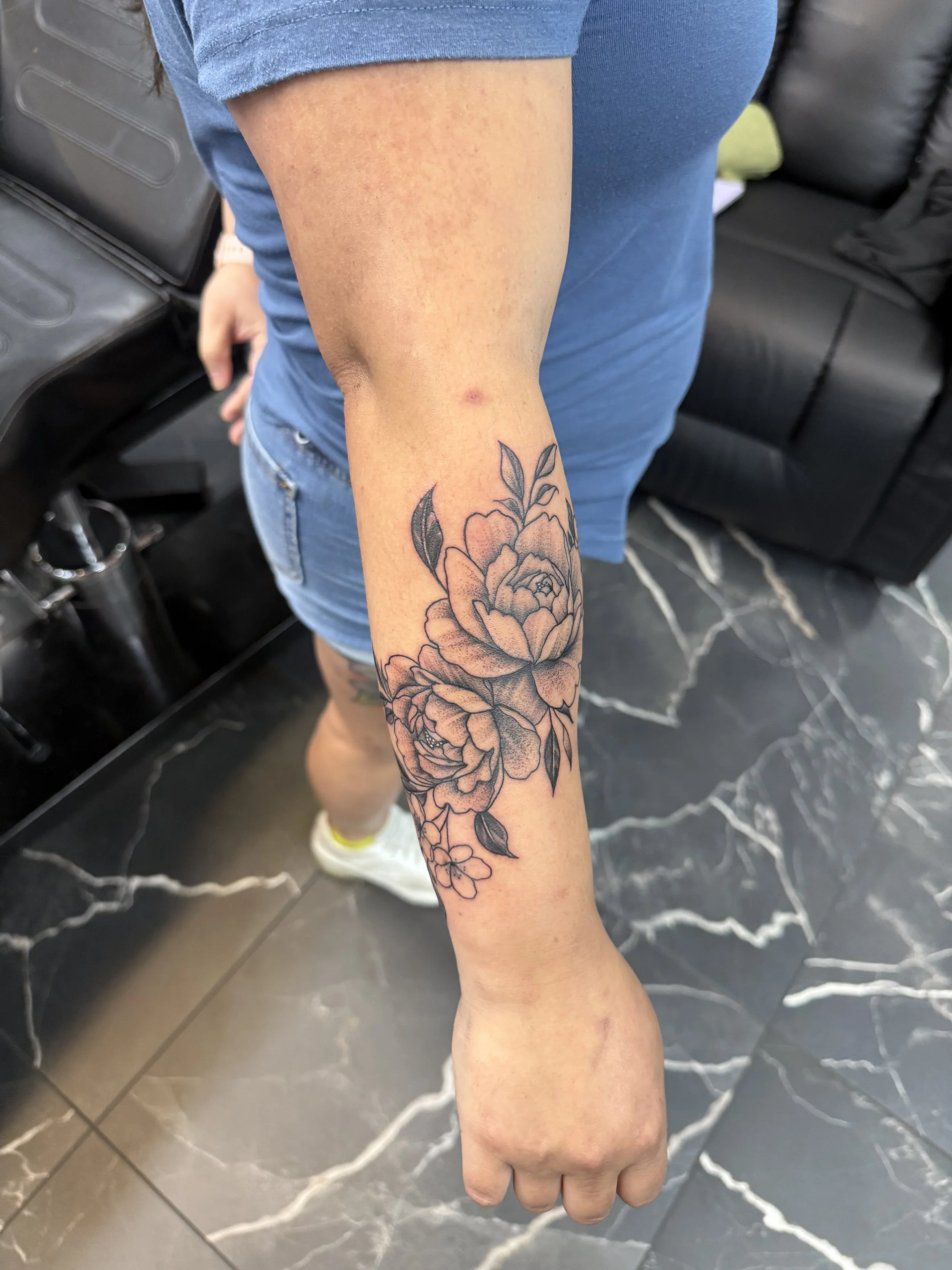 A person's arm with a floral tattoo featuring roses and leaves, wearing a blue t-shirt and blue shorts, standing on a black marble floor.