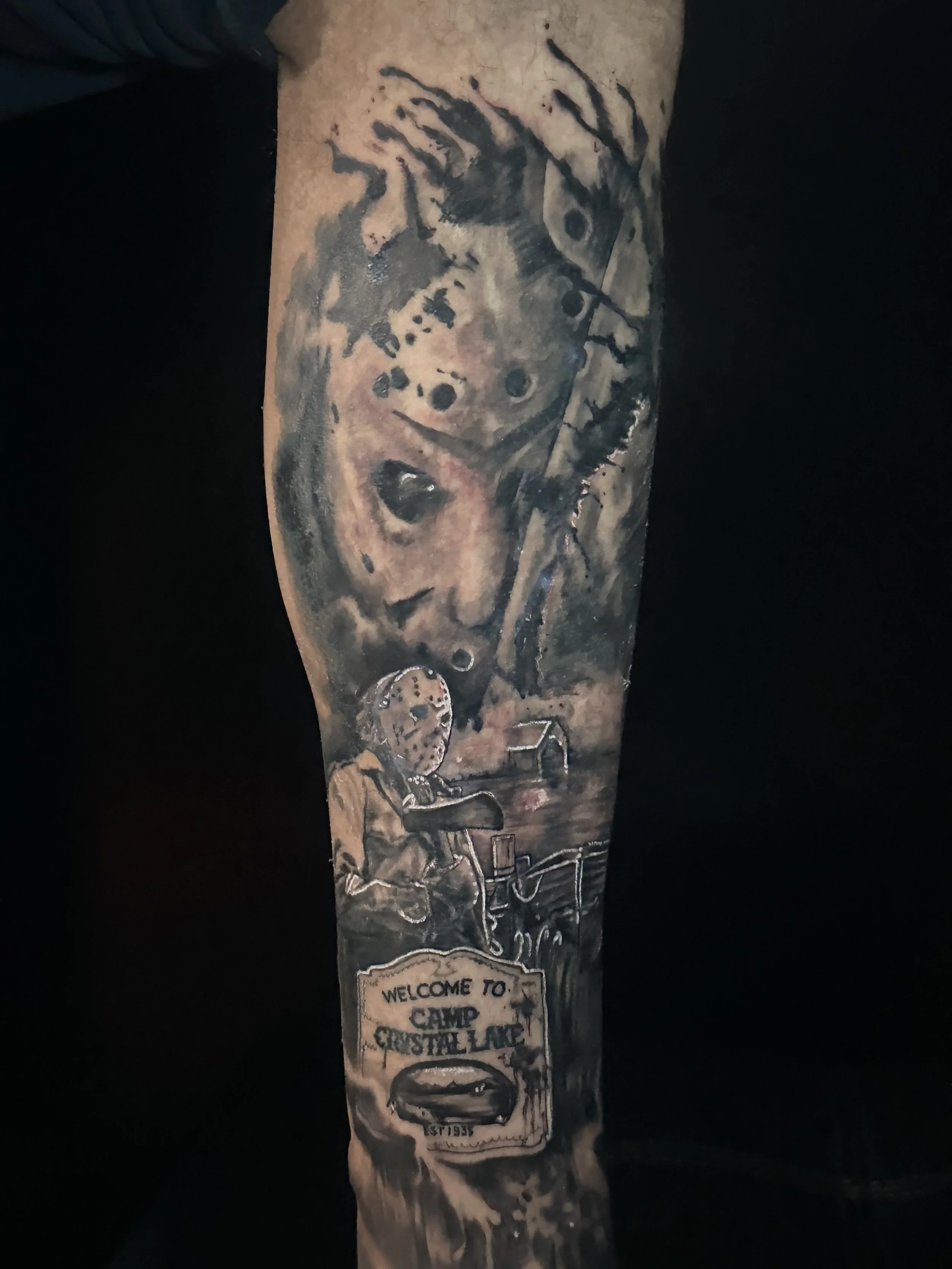Tattooed arm with various black and grey designs including a space-themed image, a circular emblem, and a sign that says "Welcome to Camp Crystal Lake."