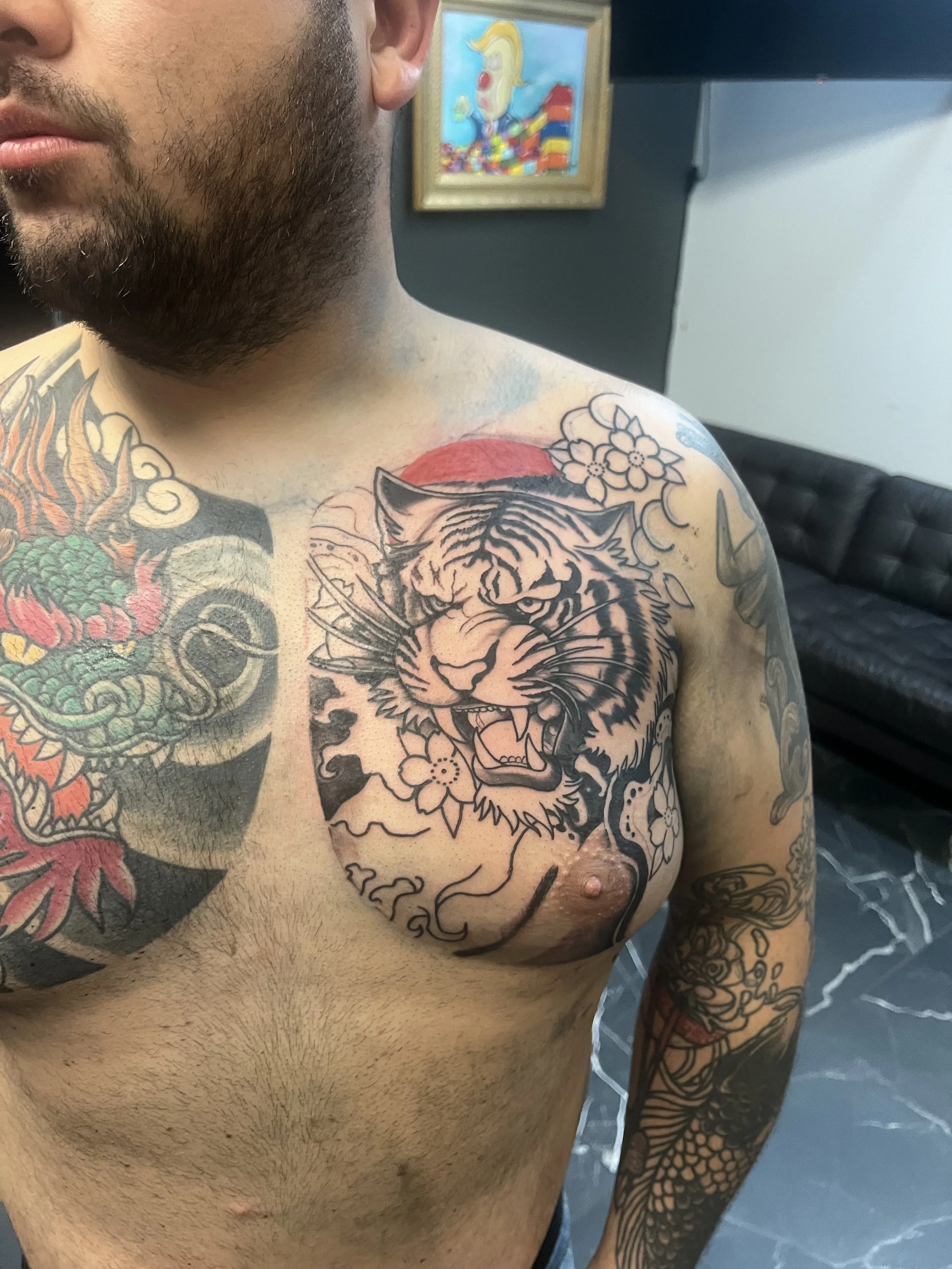 Close-up of a tattooed man shirtless, showing a tiger tattoo on his chest, a colorful dragon tattoo on his shoulder, and a sleeve tattoo on his arm.