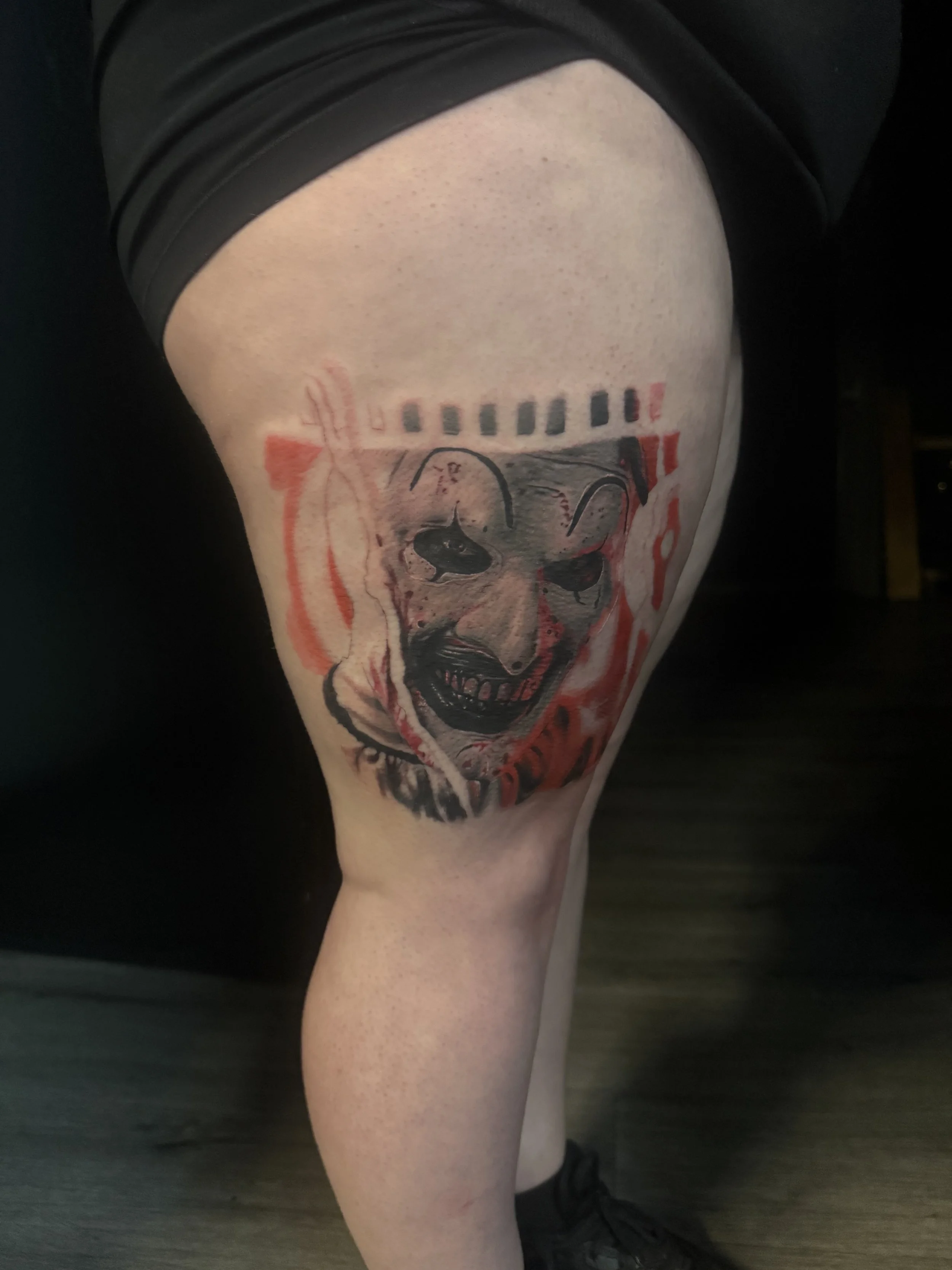 A person's thigh with a tattoo of a dog's face, wearing a black shirt and black sneakers, standing on a dark wooden floor.