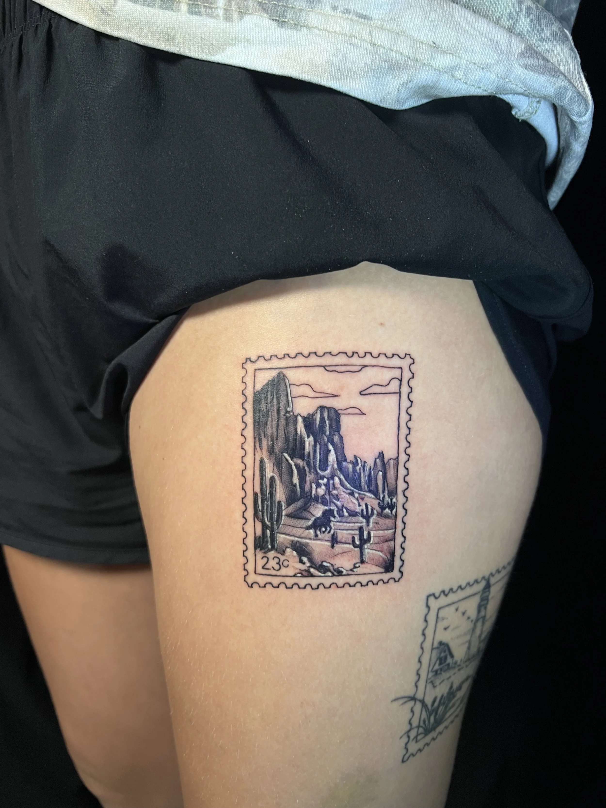 Tattoo of a scenic desert landscape with mountains, cacti, and a sunset inside a stamp-shaped border.