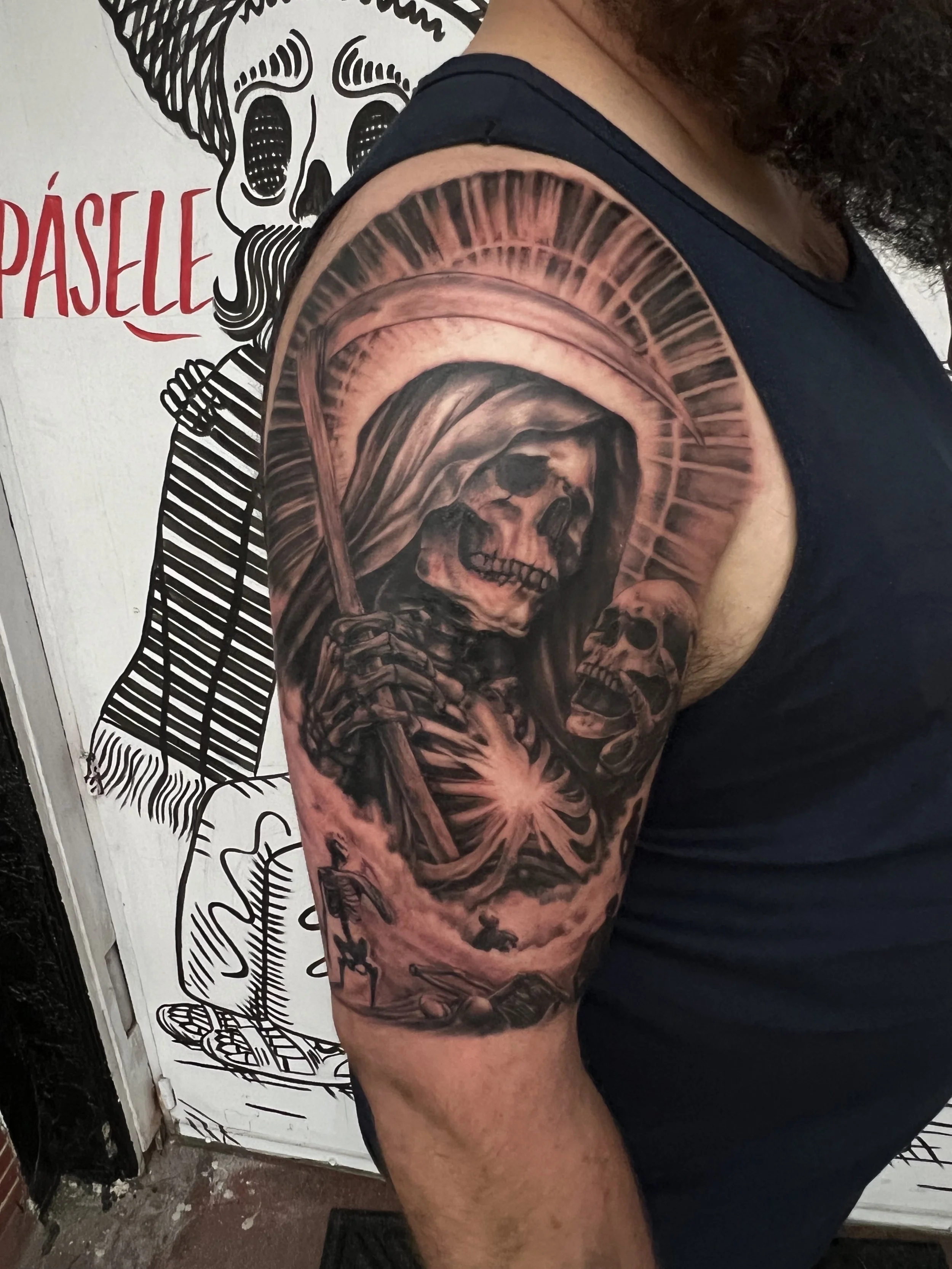 Close-up of a person's upper arm tattoo featuring a skull with a hood, surrounded by radiating lines and flames, in black and gray ink.