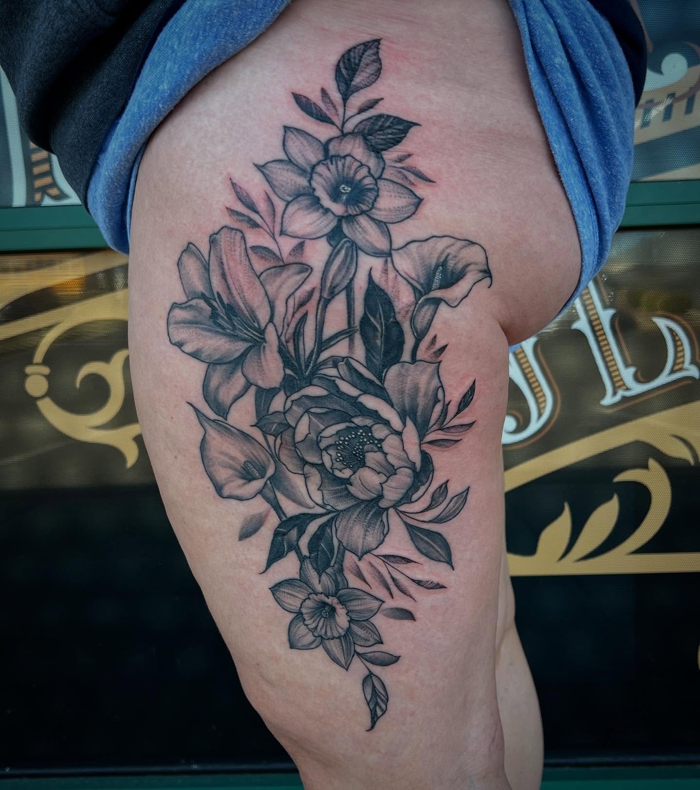 Black and gray floral tattoo on a person's upper thigh, featuring large blooming flowers, leaves, and detailed line work.